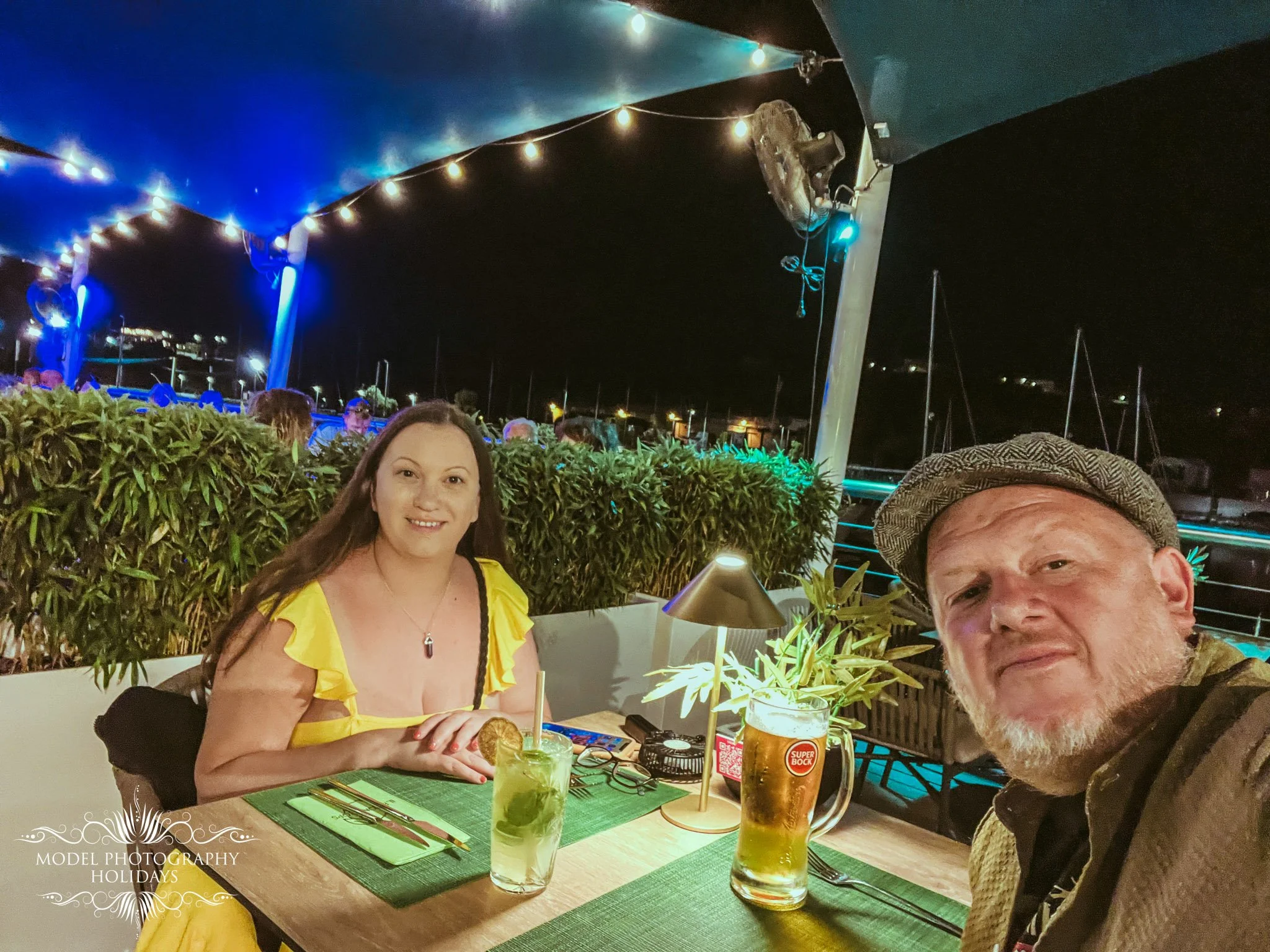 Two people dining at an outdoor restaurant at night, with drinks on the table, under string lights. One woman is smiling and wearing a yellow dress, and a man wearing a hat is partially visible in the foreground.