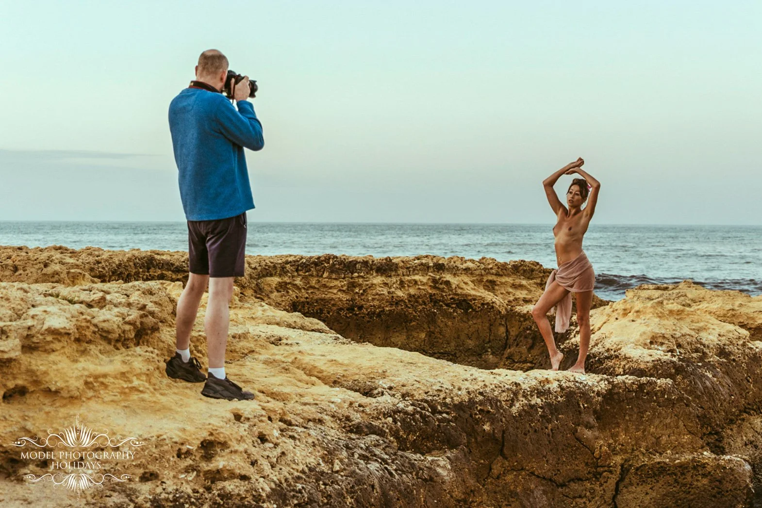 A photographer taking a picture of a woman standing on rocky terrain by the ocean, with the woman striking a pose with her arms raised above her head.