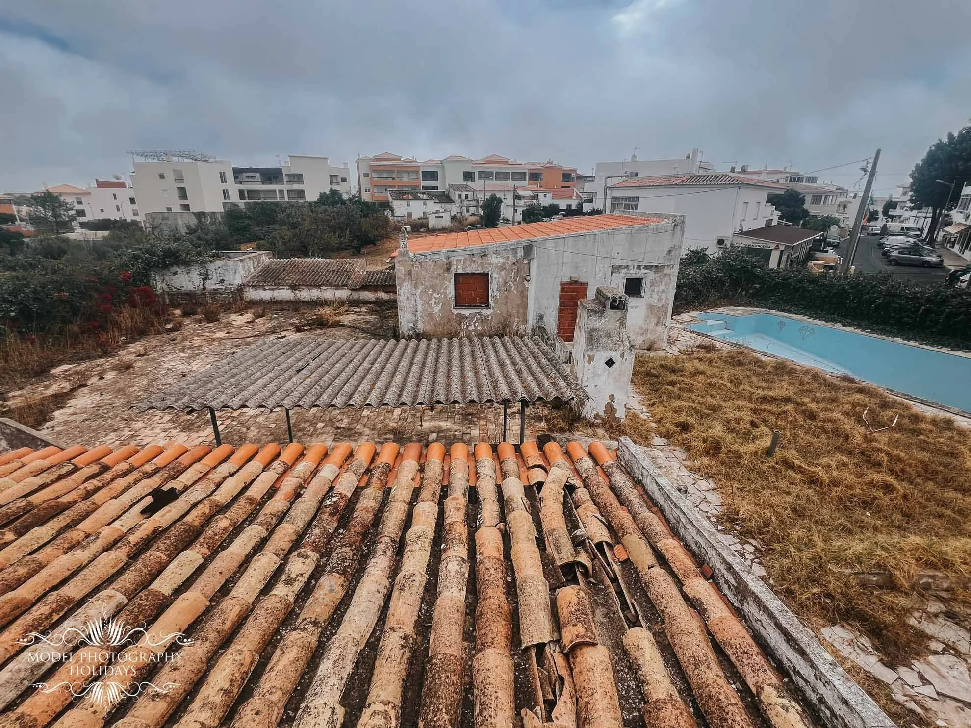 View of an old, abandoned building with a tiled roof and a neglected, overgrown yard. A pool is visible in the background. The scene is overcast with residential buildings in the distance.