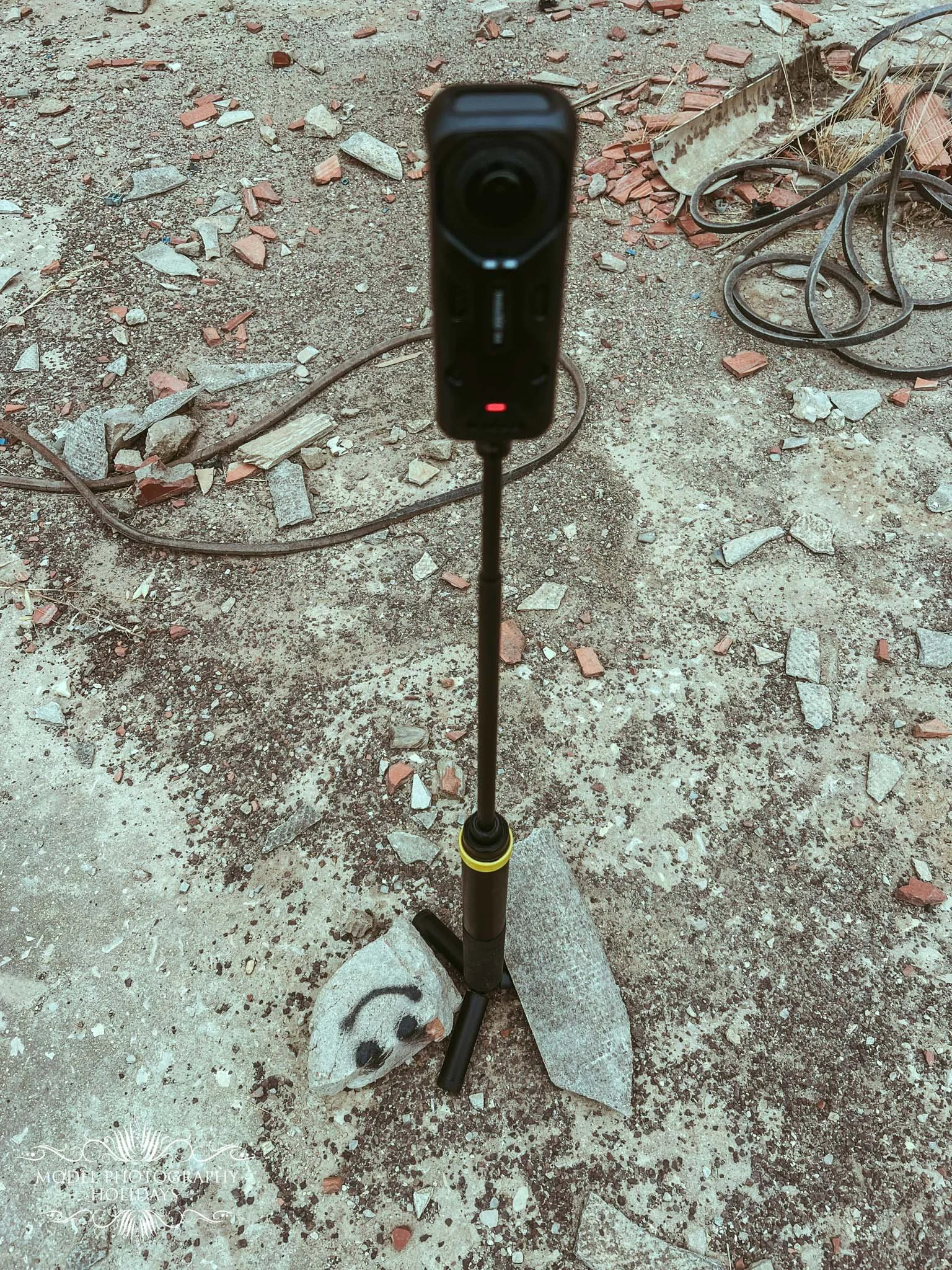 A selfie stick with a camera attached, standing on a rough dirt ground with scattered rocks, wires, and debris.