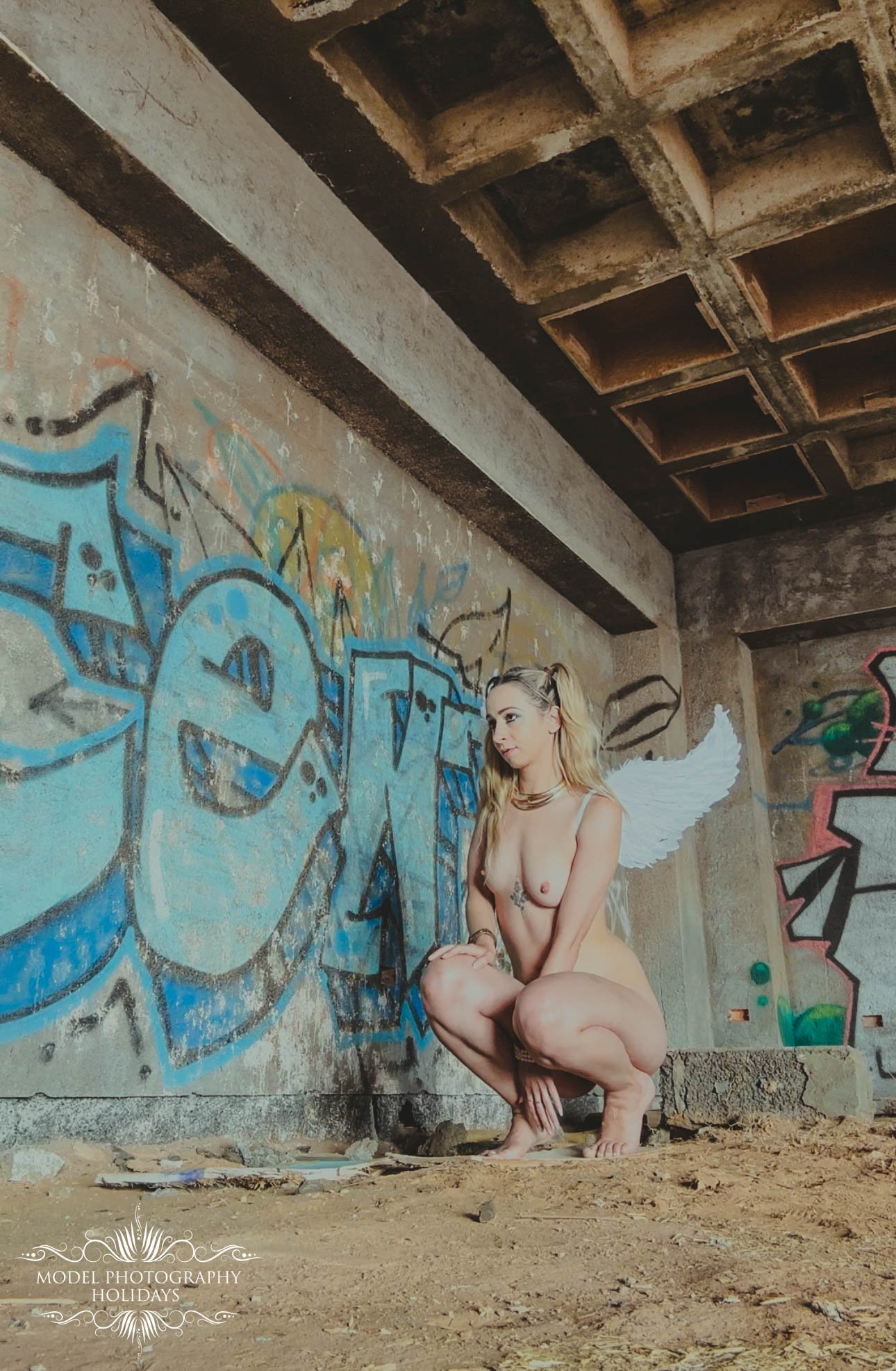 A nude model with angel wings crouching in front of graffiti on a concrete wall inside an abandoned building.