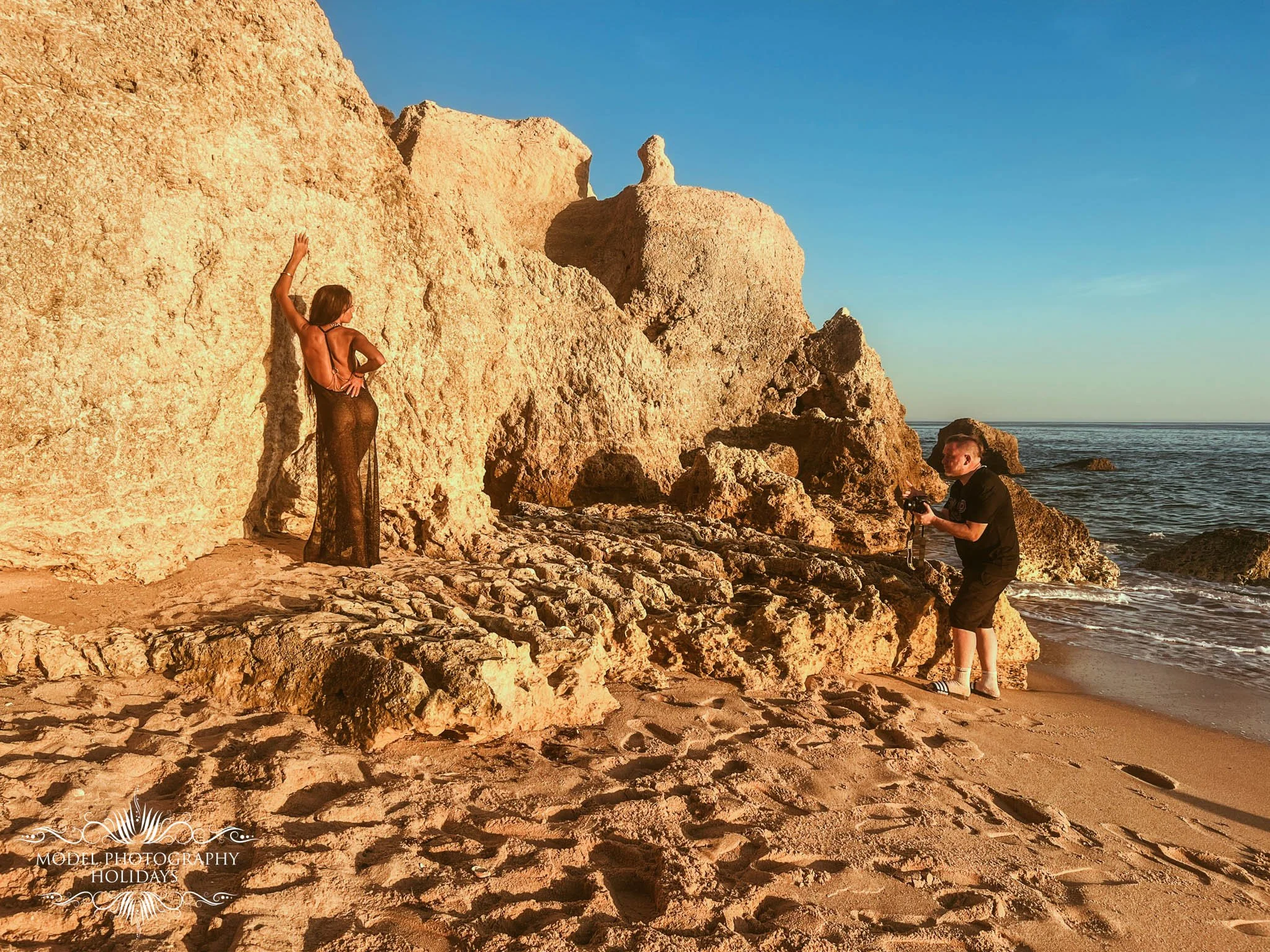 A woman with long hair wearing a see-through black dress, posing against large rock formations on a beach at sunset, with a man taking her photo.