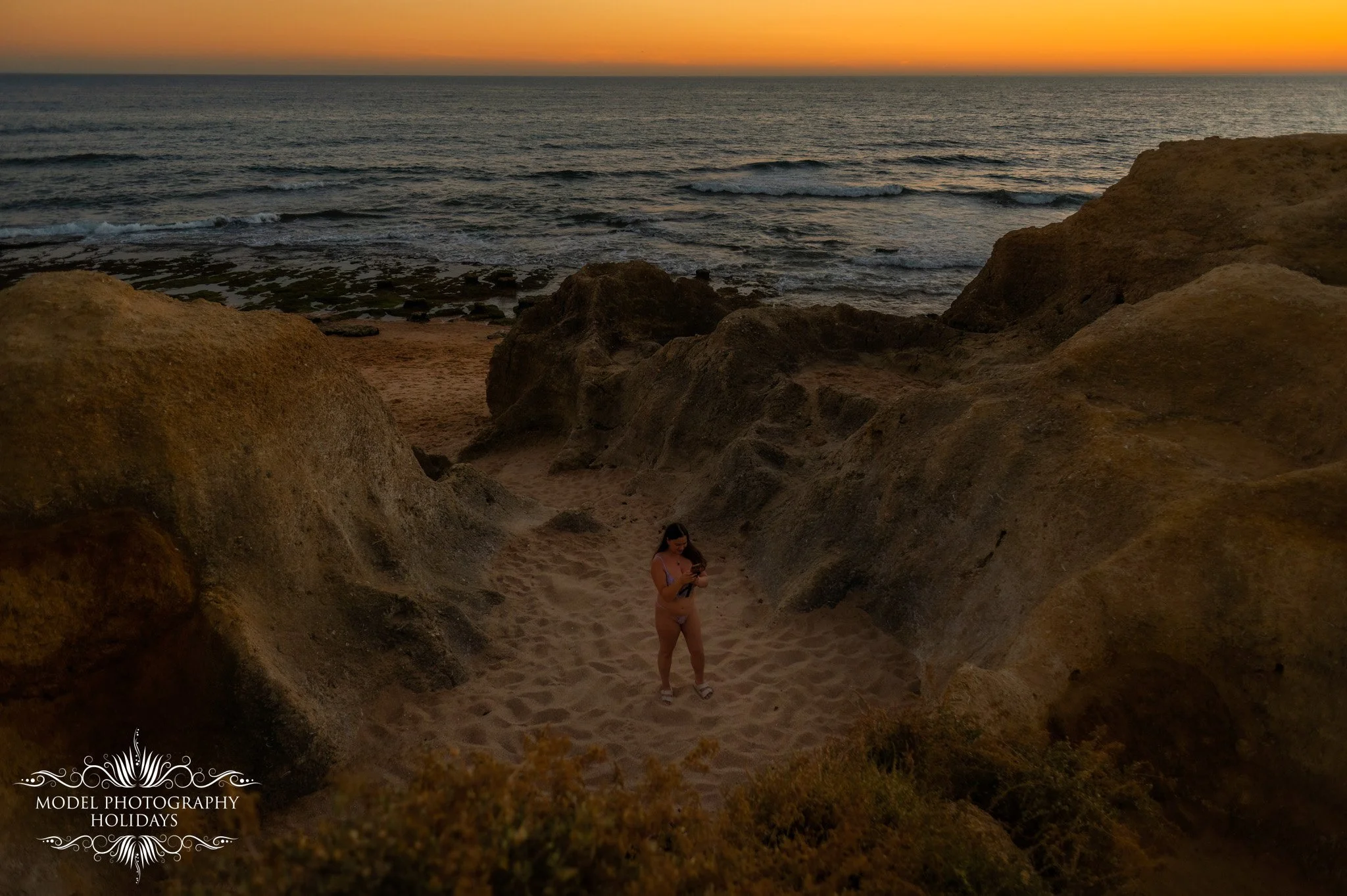 A woman in a bikini standing in a carved sandy area between large rocks, with the ocean and sunset in the background.