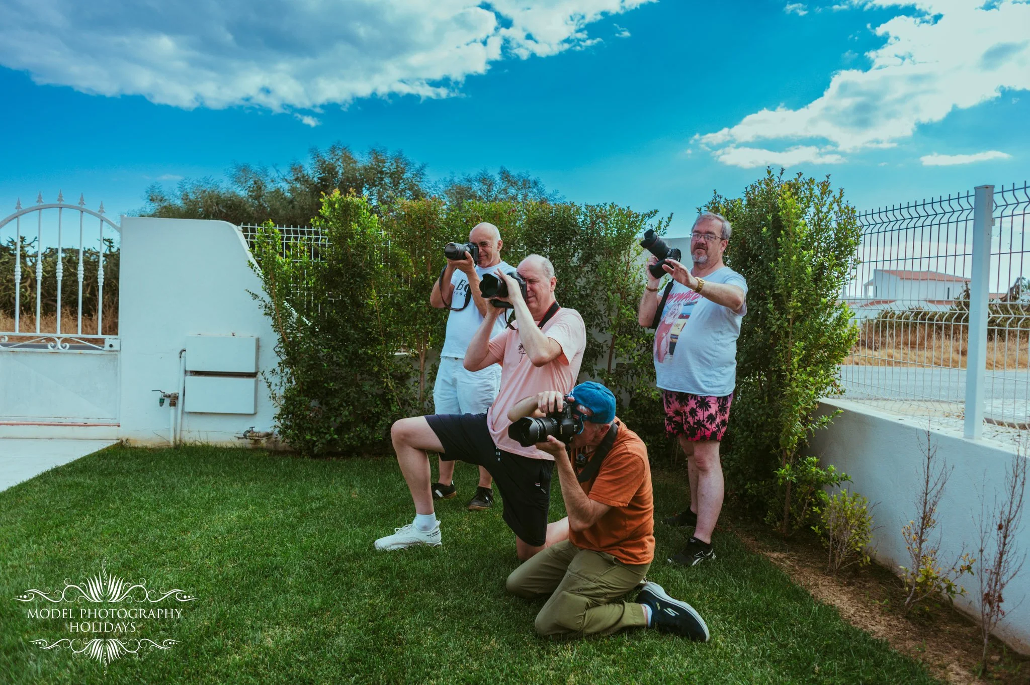 Four men, three standing and one kneeling, taking pictures outdoors with cameras. The men are on green grass in front of a white wall with a gate, bushes, and trees under a partly cloudy blue sky.