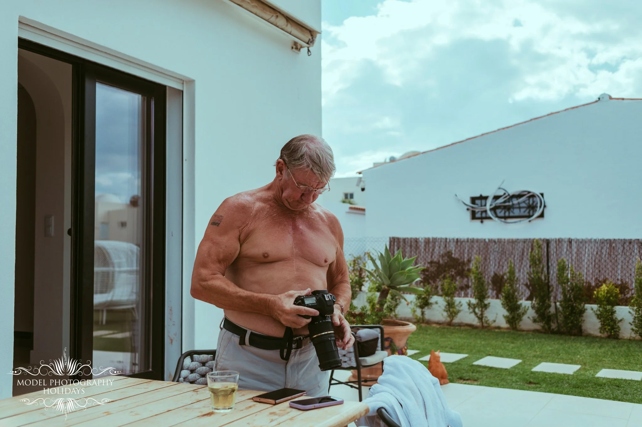 An older man with glasses and gray hair, shirtless, holding a DSLR camera, standing outside in a backyard during daytime. There is a table with a drink, a smartphone, and some clothes on it. In the background, there are plants, a fence, and a white b