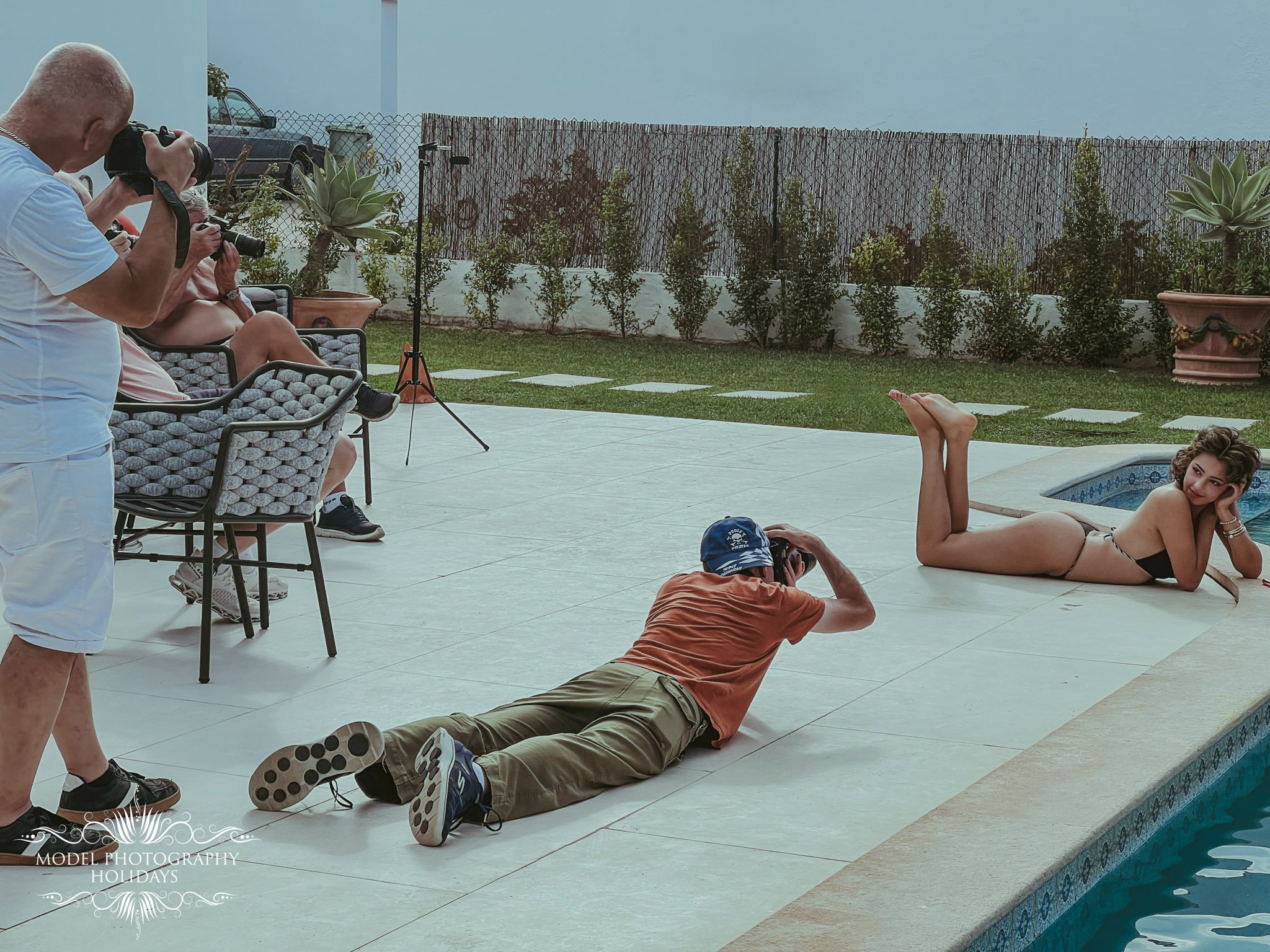 A group of people by a swimming pool, with one person lying down beside the pool and others taking photos. One woman is lying on her side in the pool, while a man is lying on the ground with a camera.
