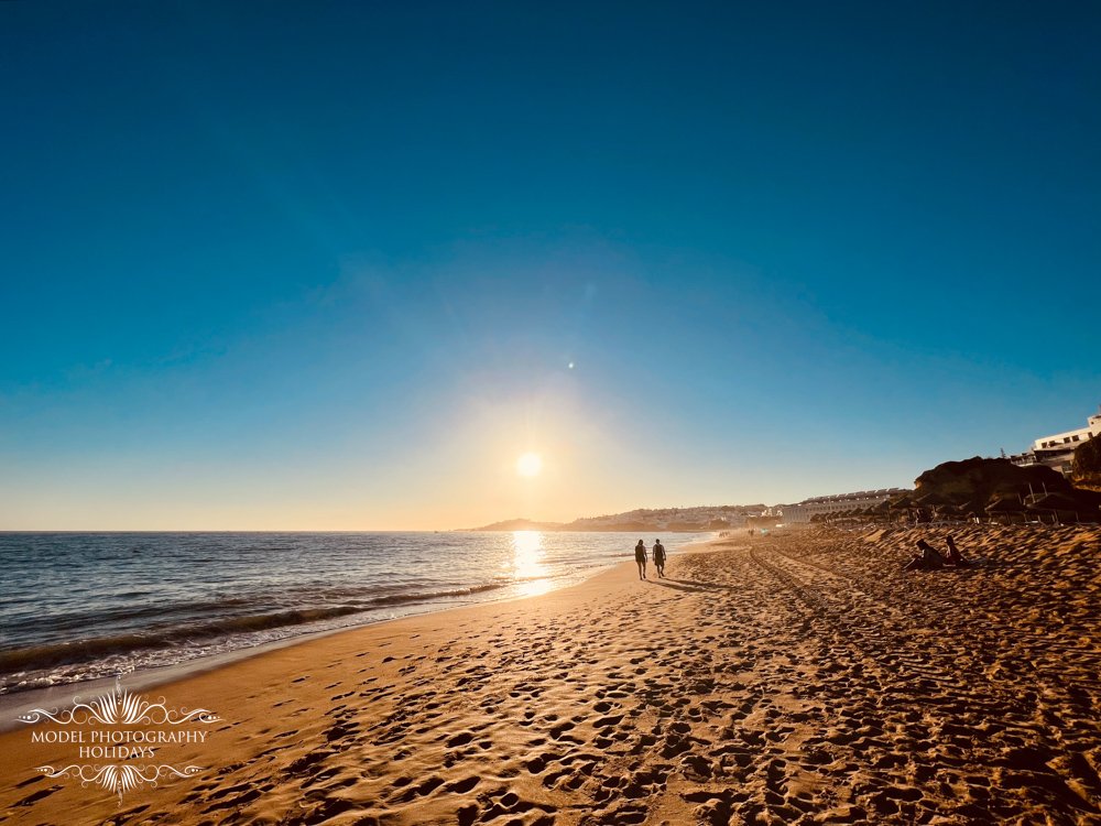 Beach sunset with people walking on sandy shore, calm ocean waves, and clear blue sky.