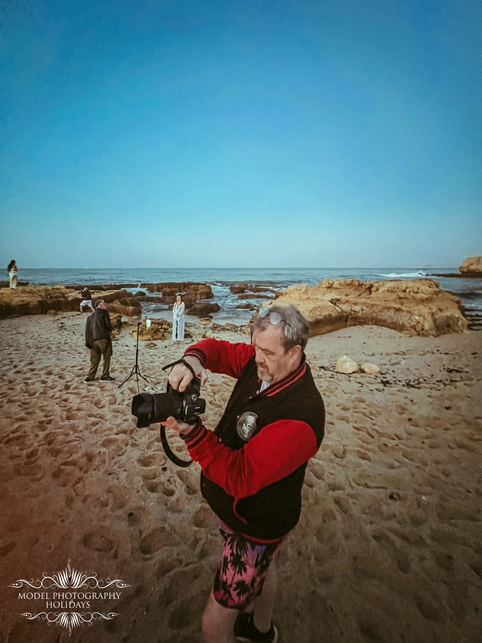 Man with gray hair and beard wearing a red and black jacket, playing with a camera on a sandy beach at sunset, with people and rocks in the background.