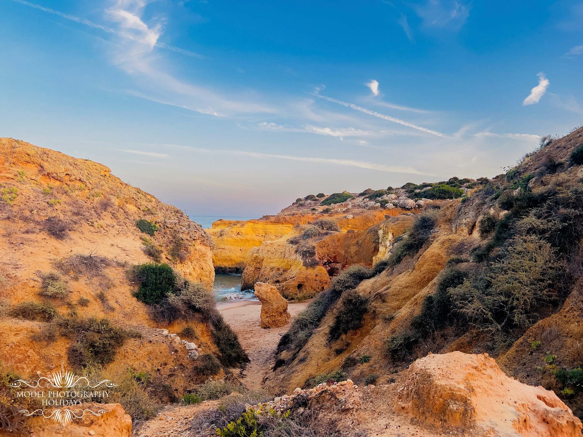 Scenic view of rocky cliffs and a narrow beach path under a blue sky with wispy clouds.
