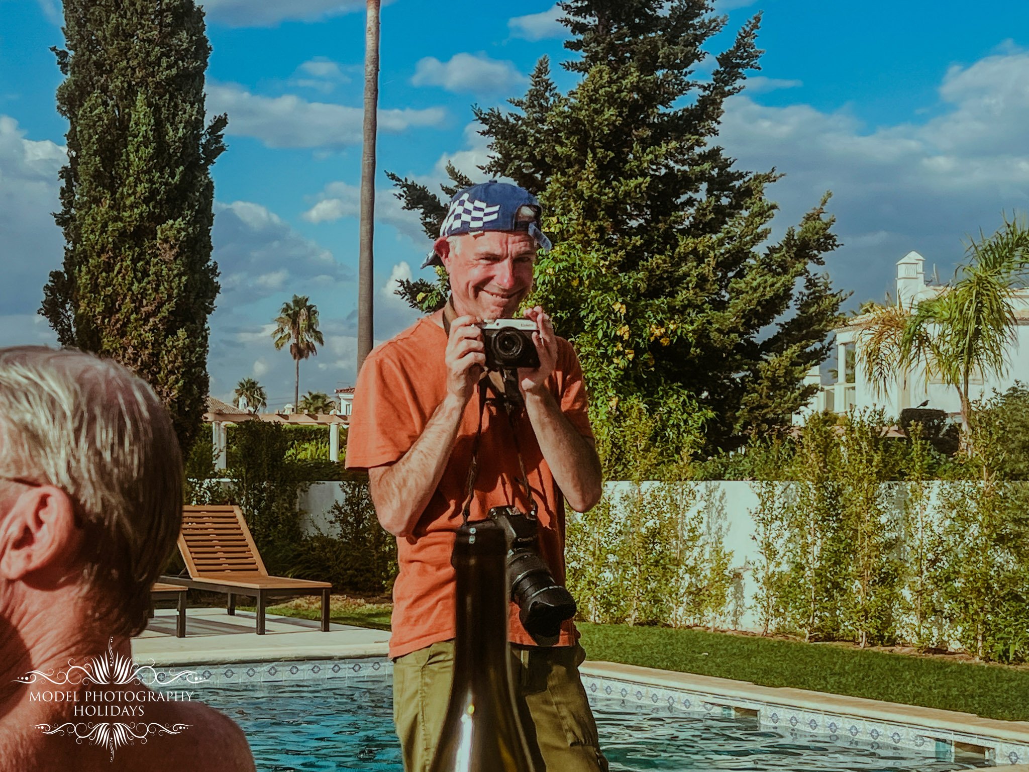 A man smiling and holding a camera, standing outdoors near a swimming pool, with trees and a blue sky in the background.