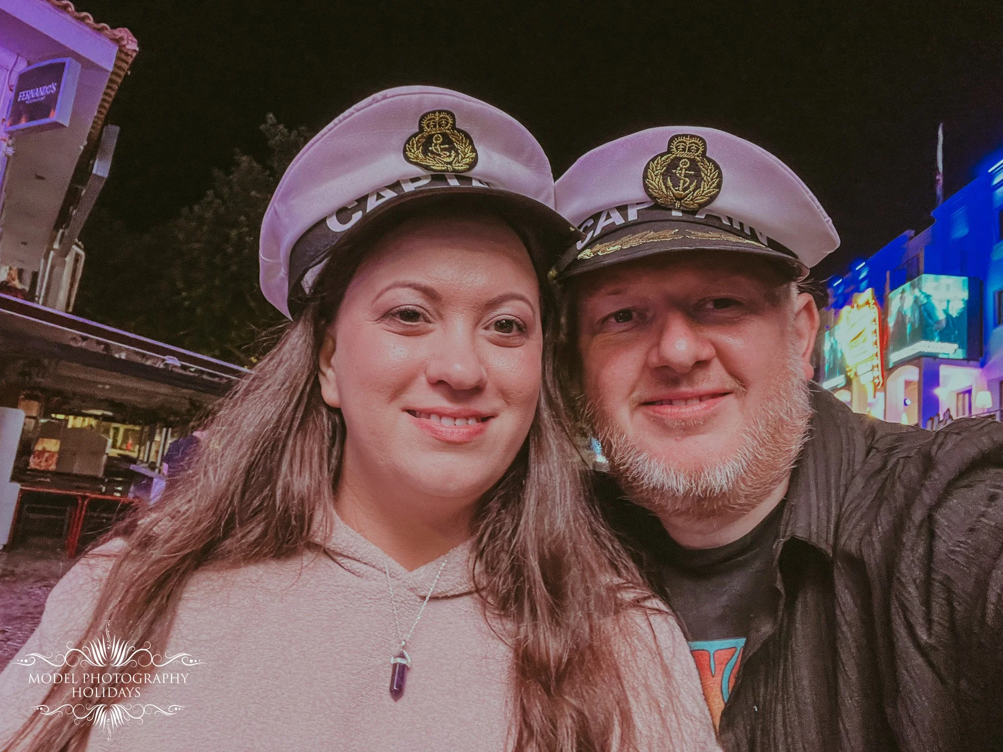 Close-up of a couple wearing captain hats at night in an urban area with bright neon lights and signage in the background.