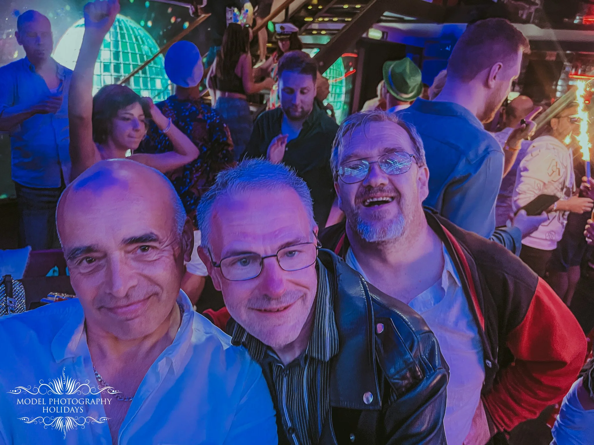 A group of people at a lively indoor event with colorful lighting. Some are dancing, chatting, and using their phones. The scene is festive with a vibrant atmosphere.