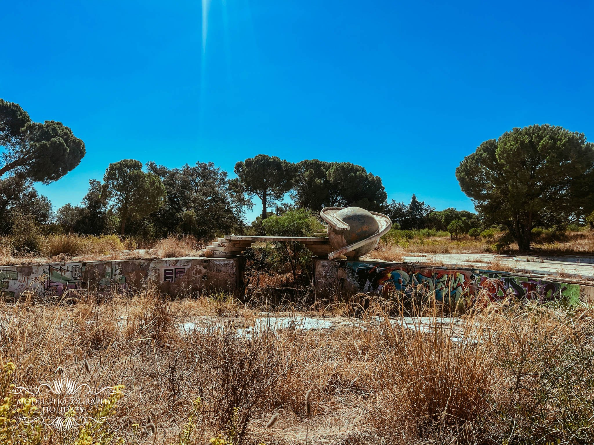 An outdoor scene featuring a large area of dry grass, graffiti-covered concrete, trees, and a large old boat set on the ground under a clear blue sky.