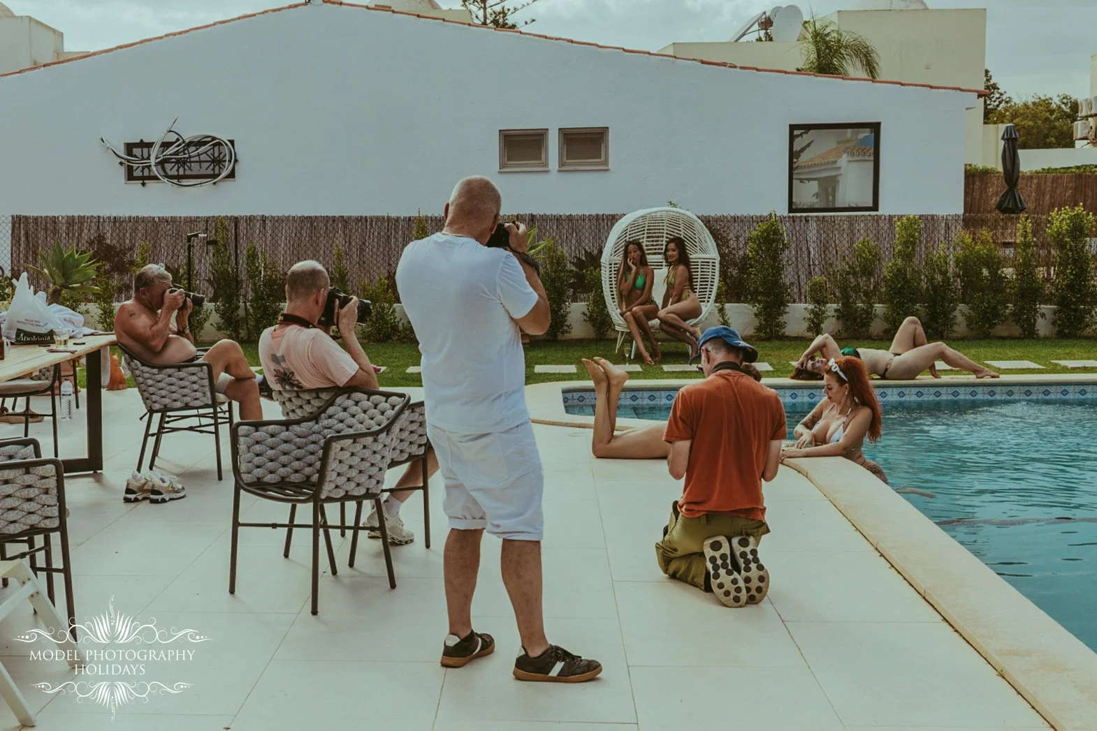 Photographers capturing models in swimsuits by a backyard pool, with some models lounging on the grass and others in the water, during a photo shoot.