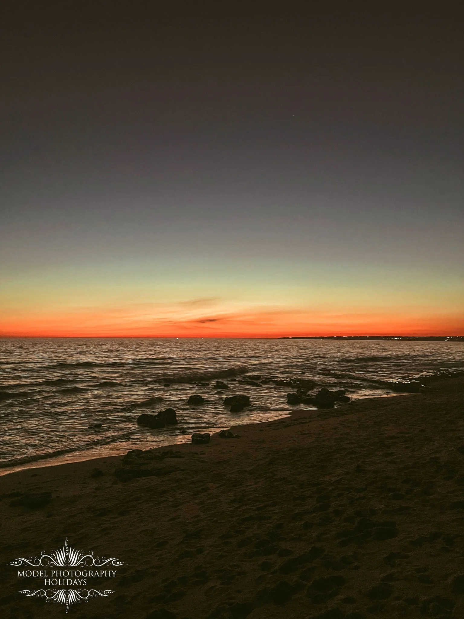 A sunset over the ocean with a colorful sky, dark waters, and a sandy beach with rocks in the foreground.