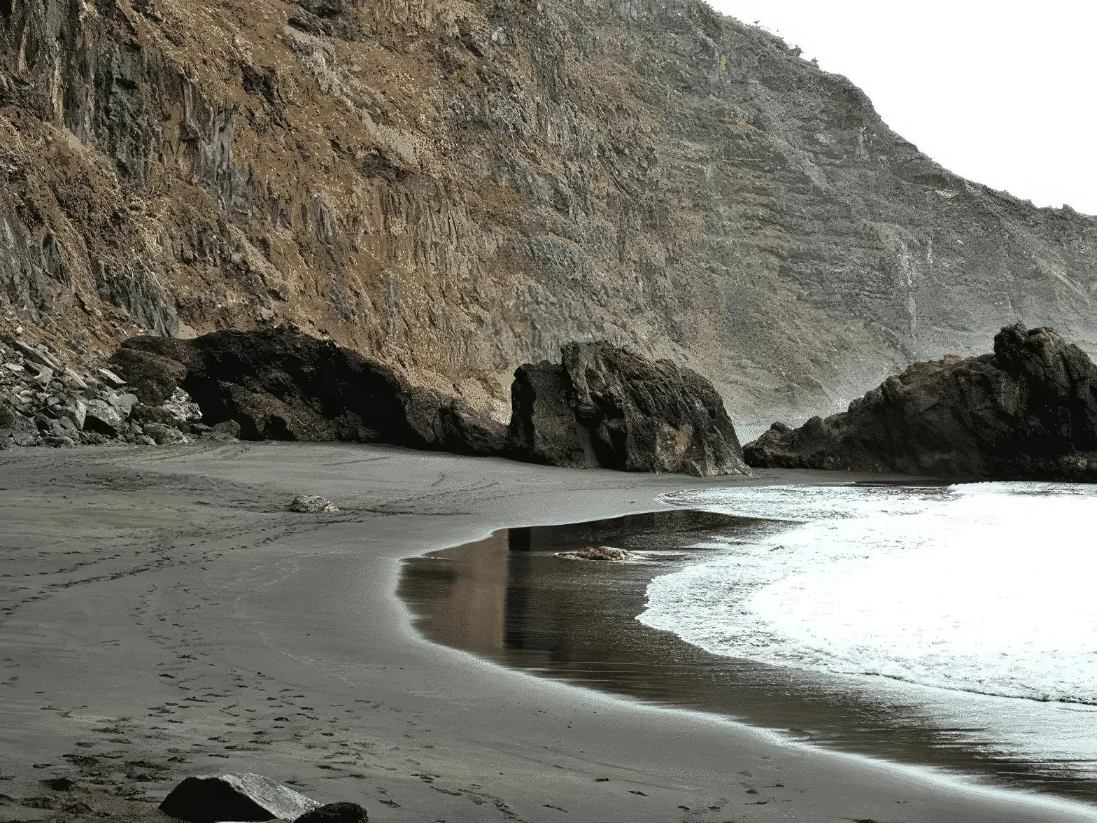 A rocky cove with dark sand, large rocks, and cliffs on one side, and gentle waves in the ocean on the other.