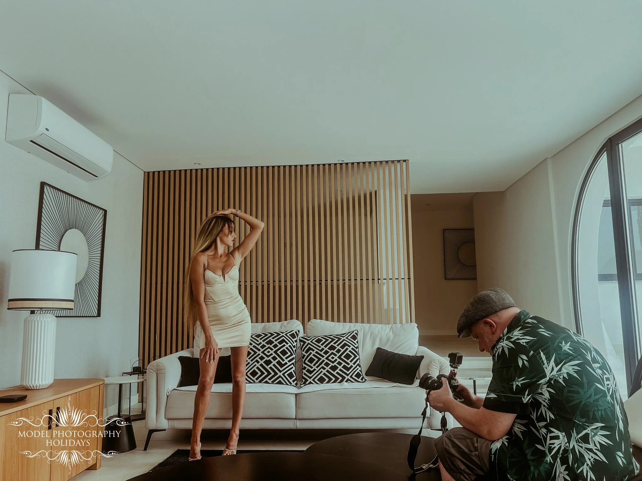 A woman in a beige dress posing for a photoshoot in a modern living room with a wooden slatted wall, a white sofa with patterned cushions, and large windows. A photographer is kneeling in front of her with a camera, capturing her image.