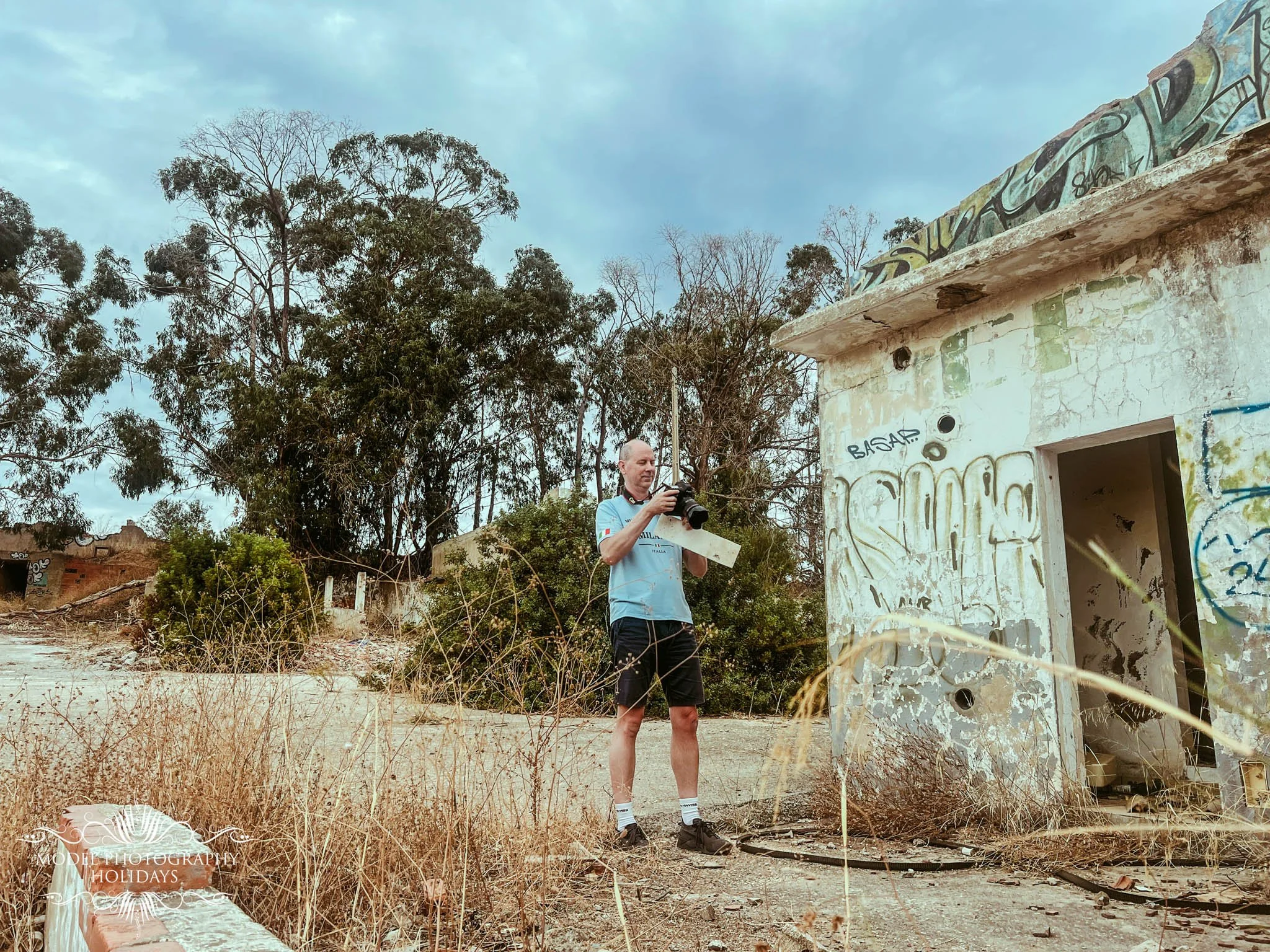 A man in a light blue cycling jersey and black shorts holds a camera, standing in front of an abandoned building covered in graffiti with a cloudy sky overhead and trees in the background.