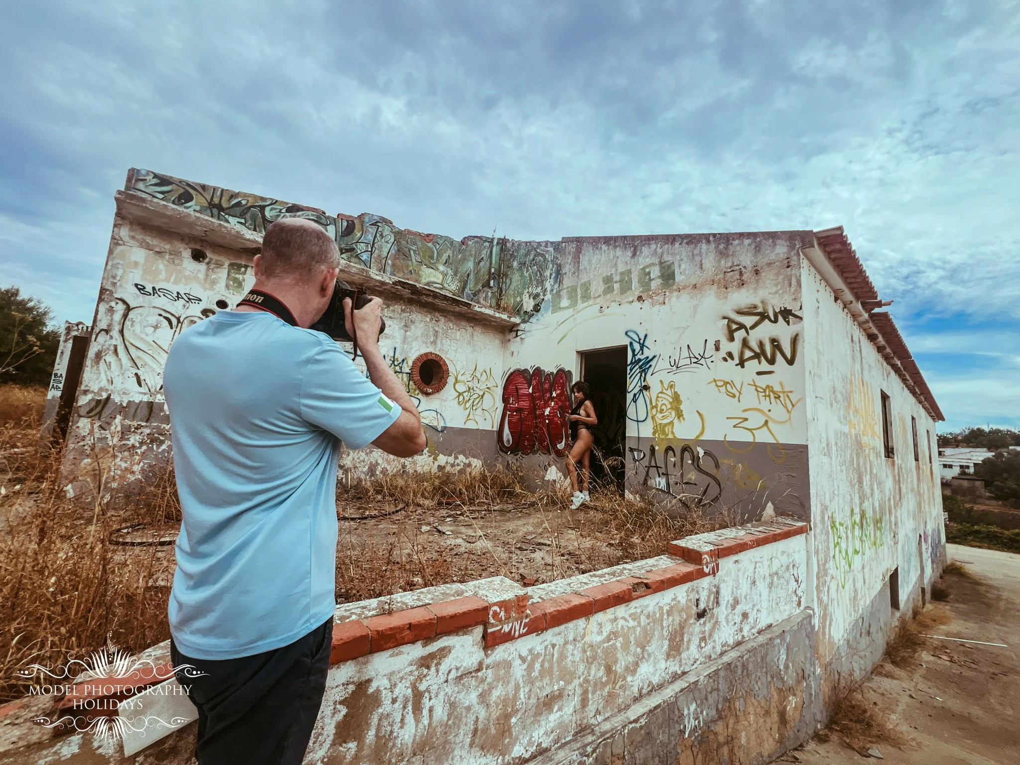 A man with a camera taking photos of a woman standing outside a graffiti-covered building with a cloudy sky overhead.