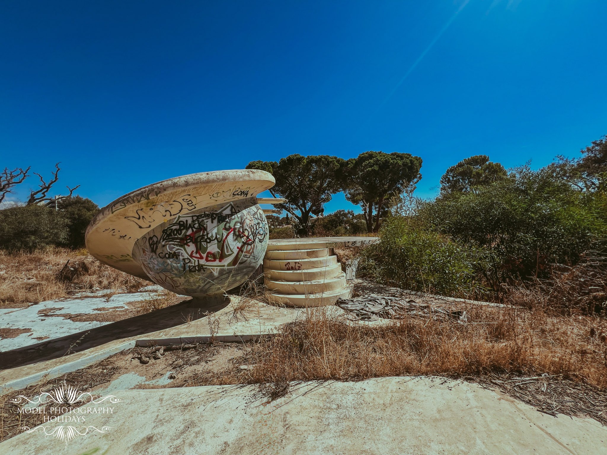 An abandoned, graffiti-covered, upside-down boat or sculpture on a concrete stand in an outdoor area with dry grass, trees, and clear blue sky.