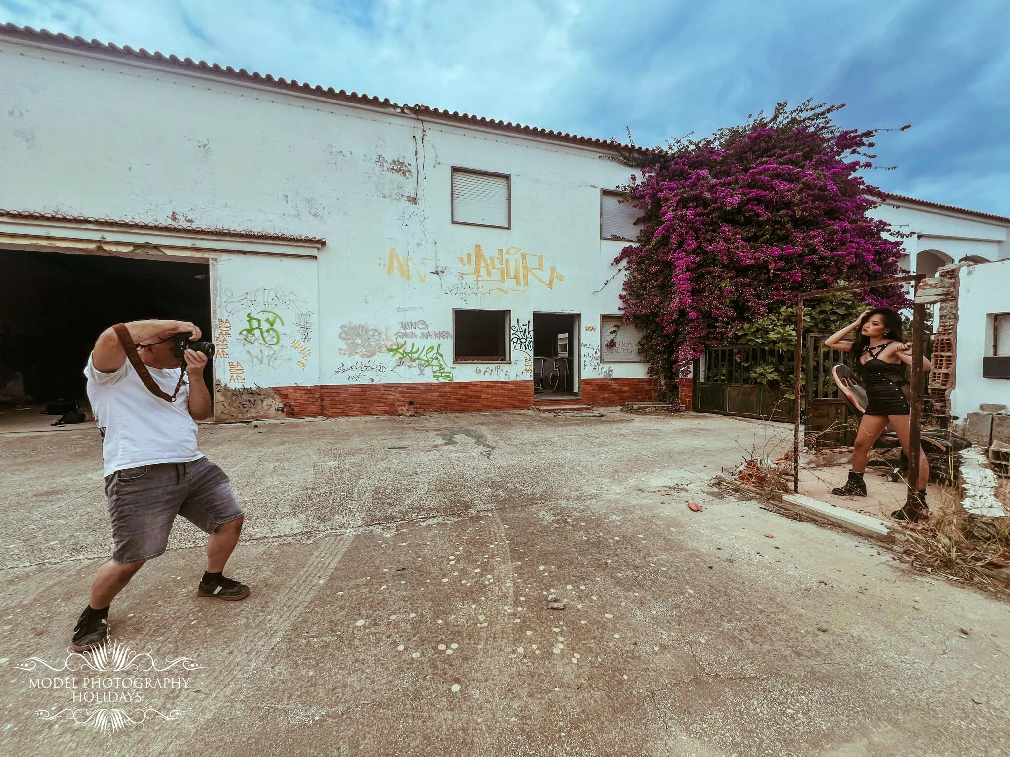 A man taking a photo of a woman sitting on a chair outside a white building with graffiti, pink flowering bushes, and cloudy sky.