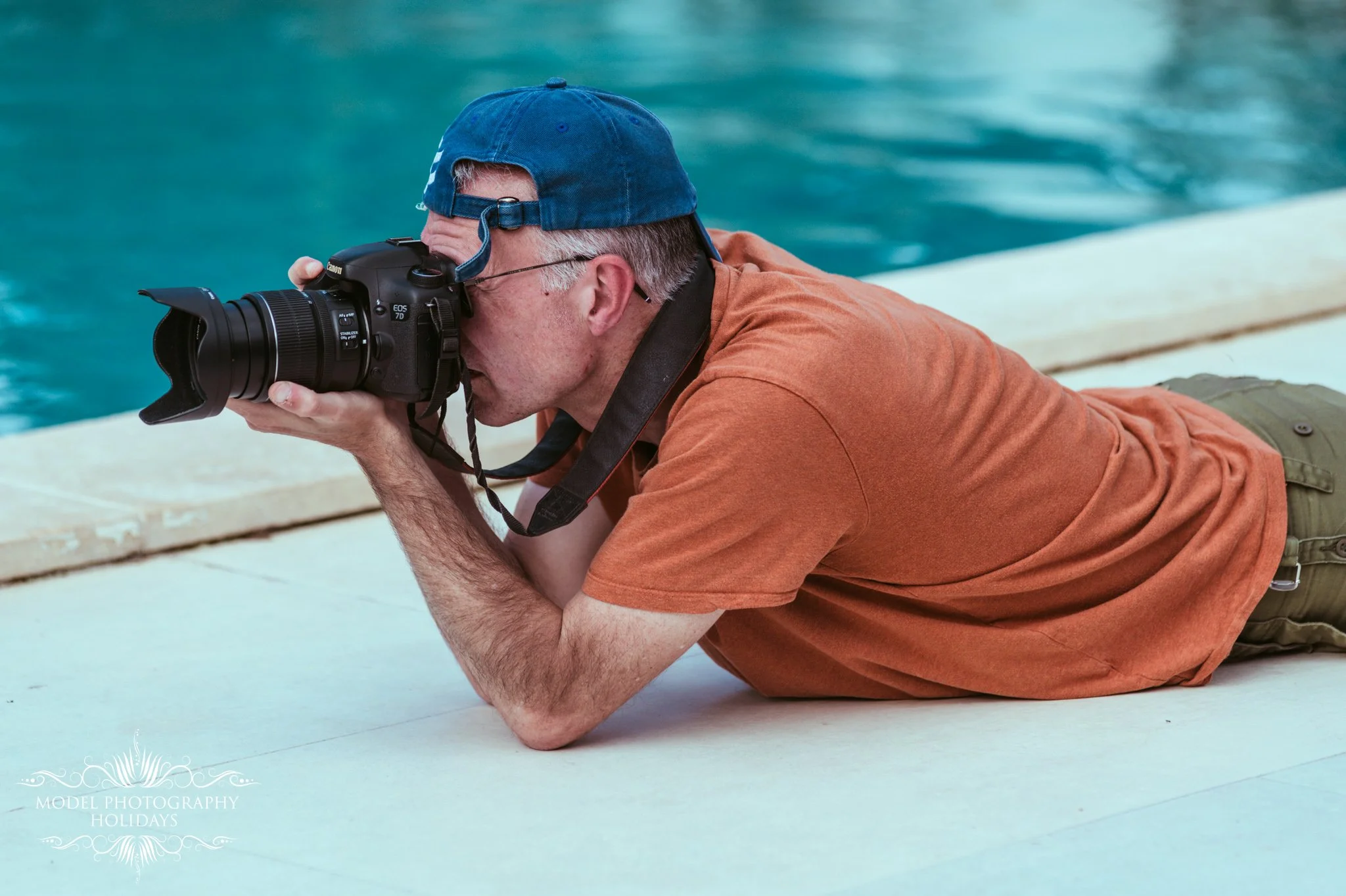 A man wearing a blue baseball cap and an orange shirt is lying on his stomach on the ground, taking a photograph with a professional camera by a swimming pool.