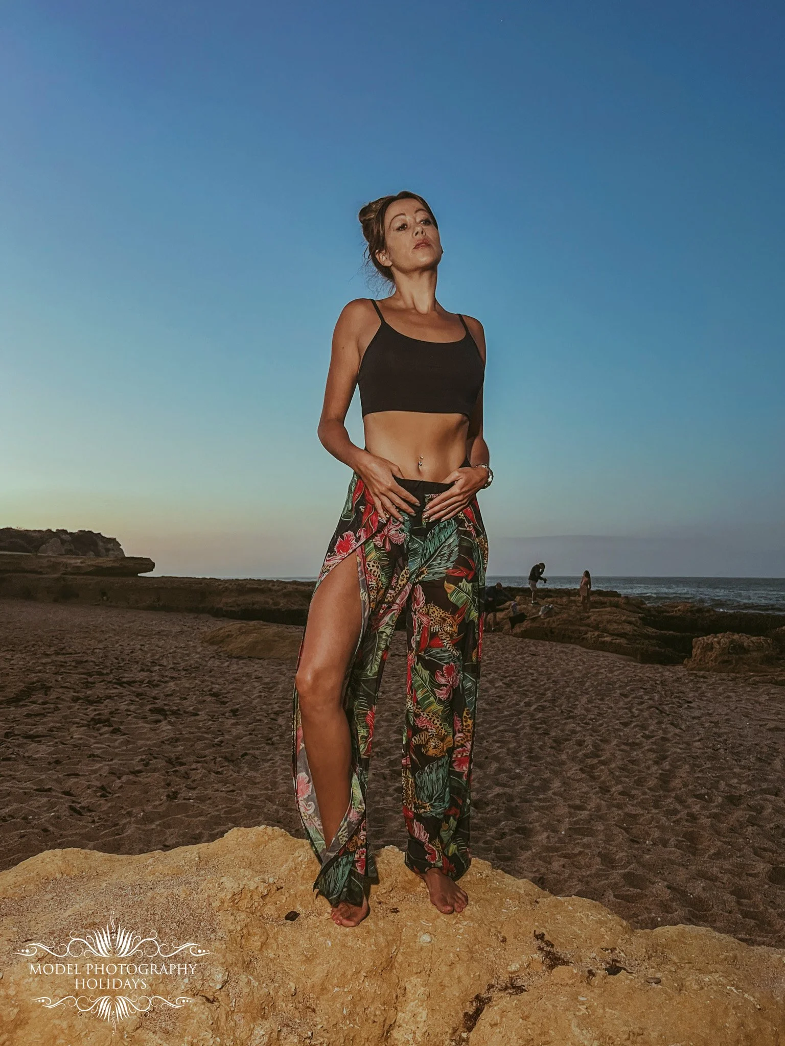 A woman standing barefoot on a rocky surface at sunset on the beach, wearing a black crop top and colorful, floral, wide-leg pants with a high slit. She has her hands near her waist, looking confidently at the camera with a serious expression. The oc