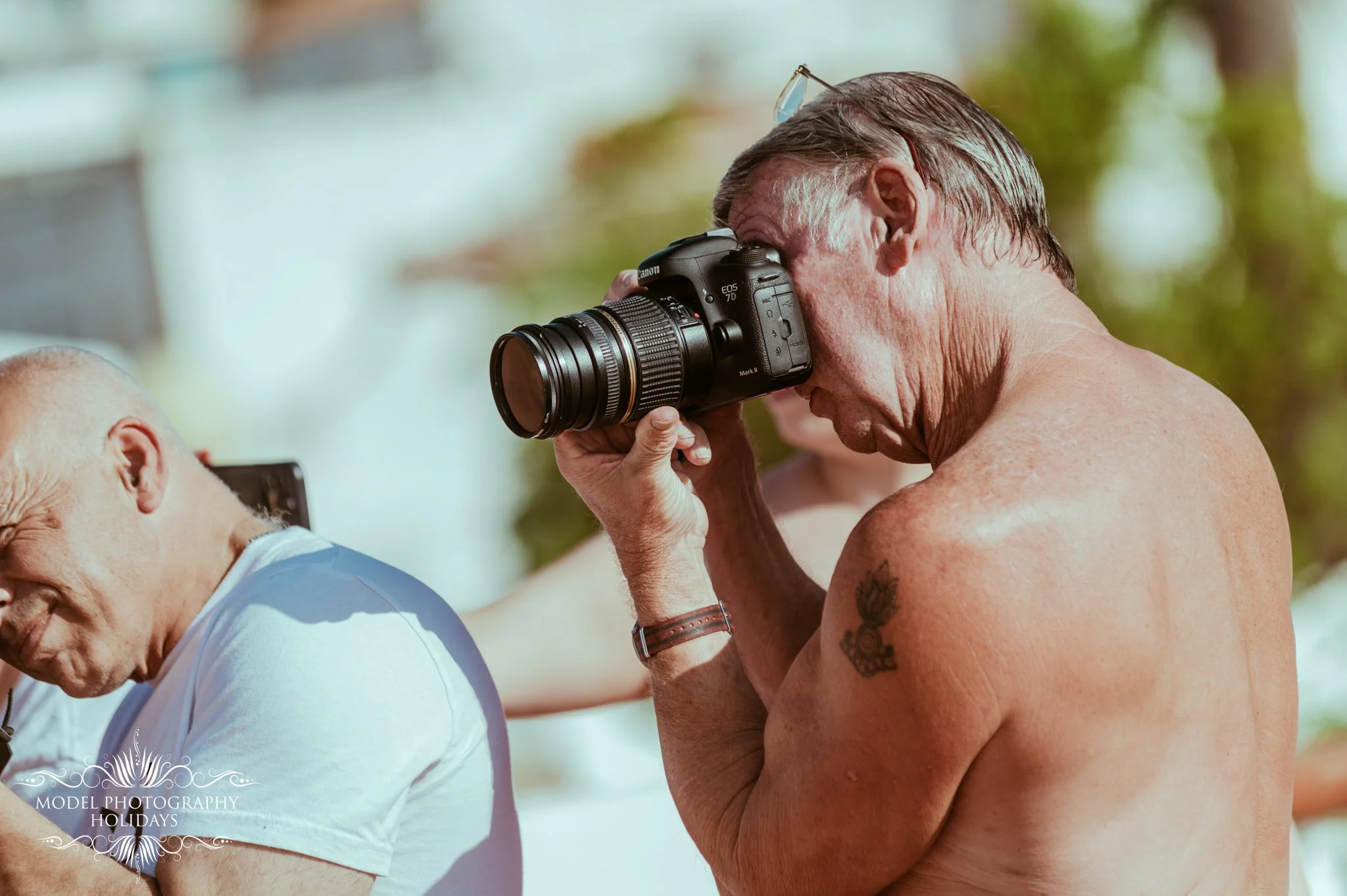 A shirtless man with glasses on his head taking a photograph of an elderly man with a camera during a sunny outdoor event.