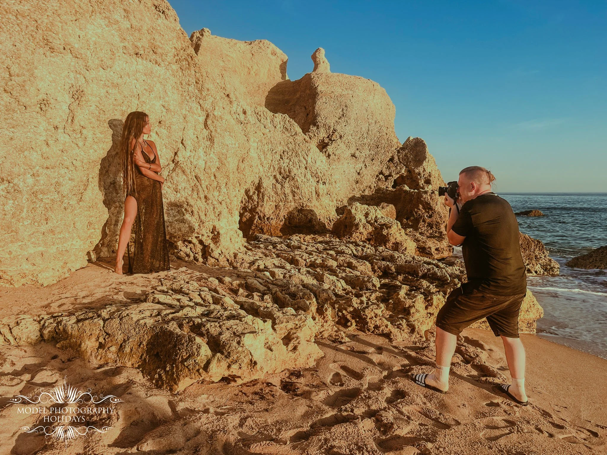 A woman in a long, sheer dress stands next to large rock formations at the beach, while a man takes a photograph of her near the shoreline with rocky cliffs in the background and the ocean on the right.