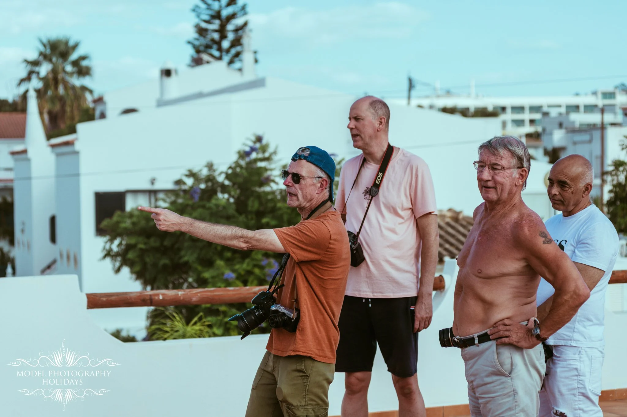 Four men walking outdoors in a warm climate, two shirtless, one wearing a hat and sunglasses, with houses and trees in the background.