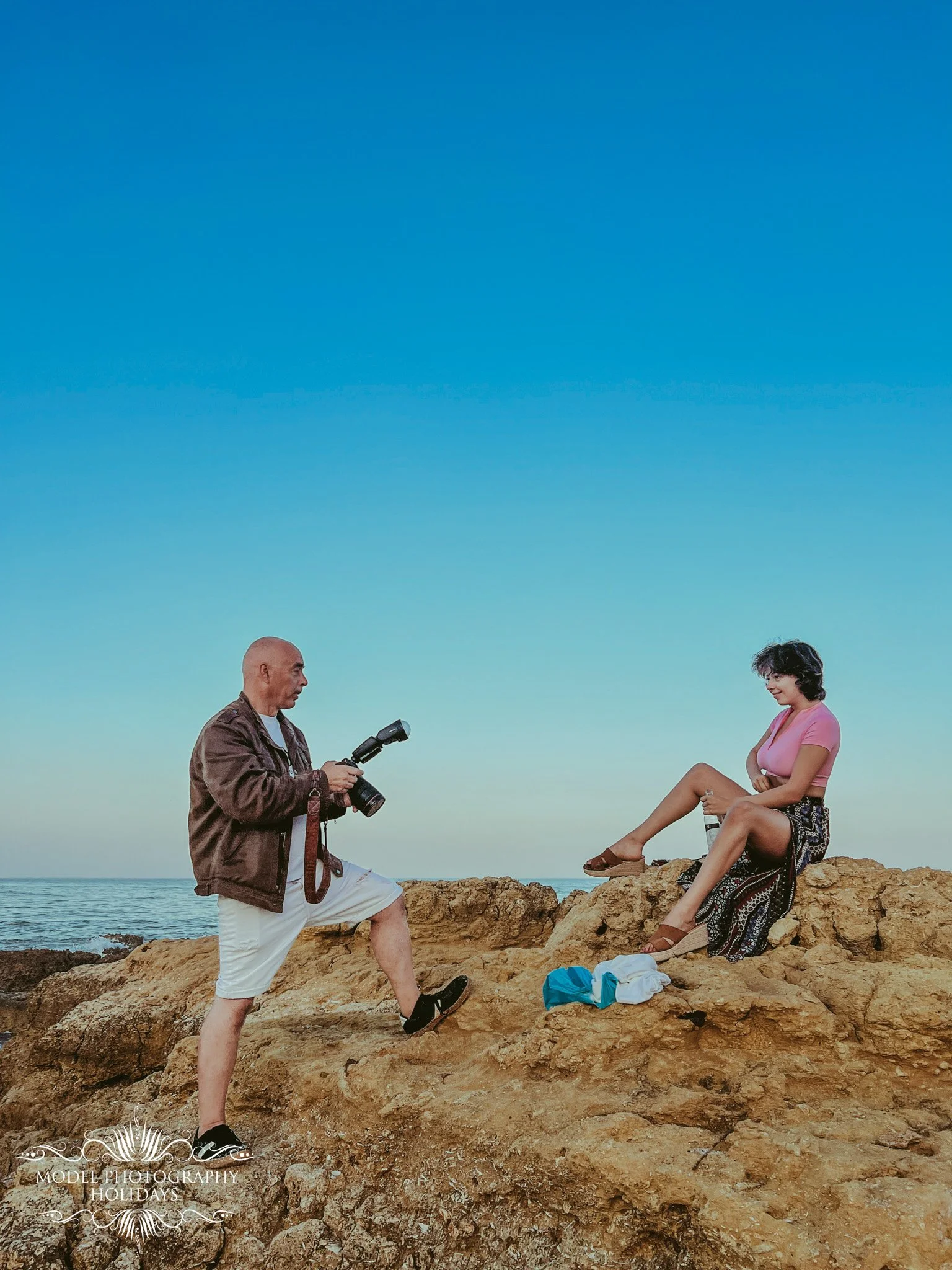 A man in a brown jacket and white shorts is taking a photo of a woman sitting on a rock by the ocean. The woman, wearing a pink top and patterned pants, is smiling, with her legs crossed and arms resting on her knee. The scene is set on a rocky coast