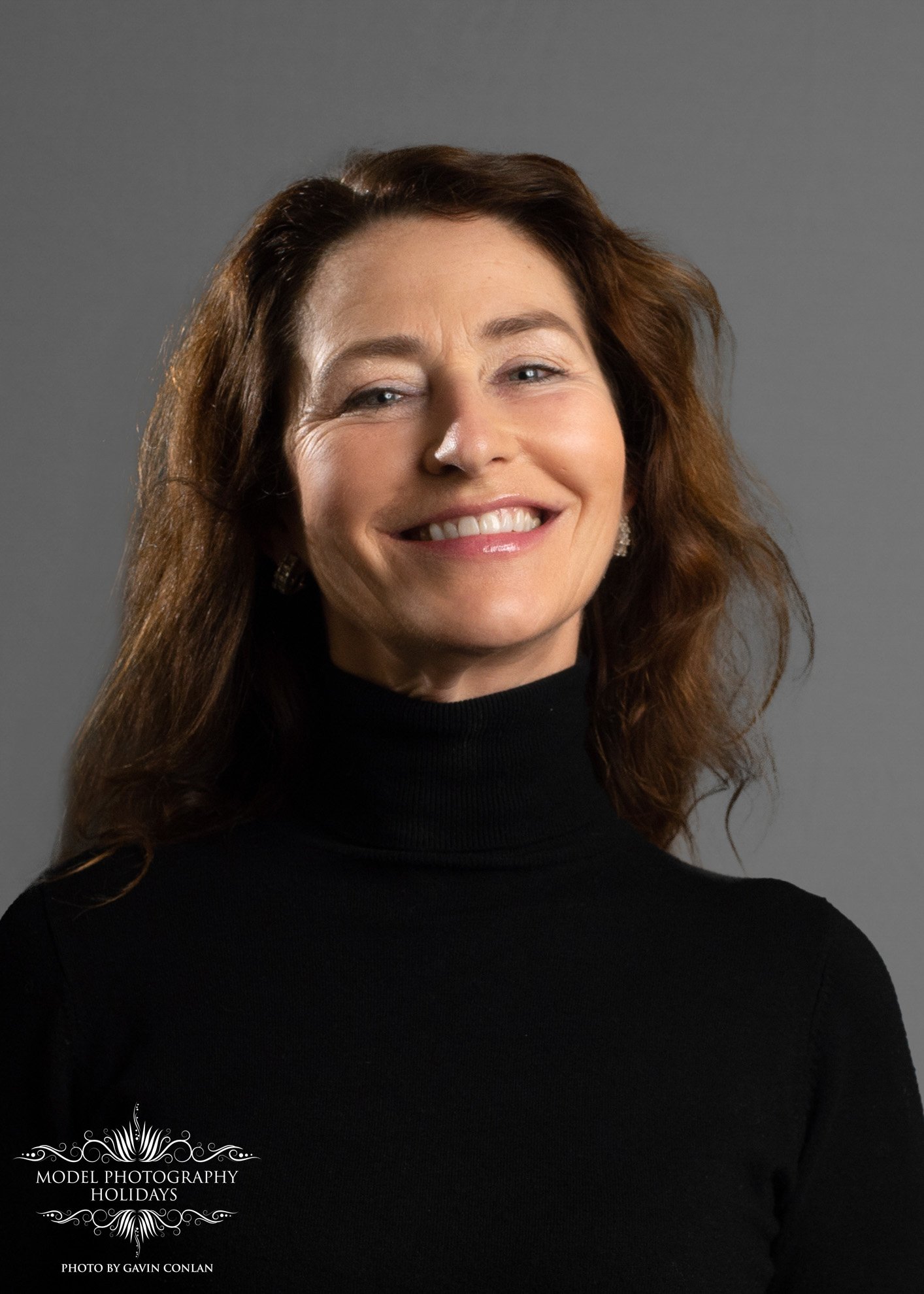 Smiling woman with long brown hair wearing a black turtleneck against a gray background.