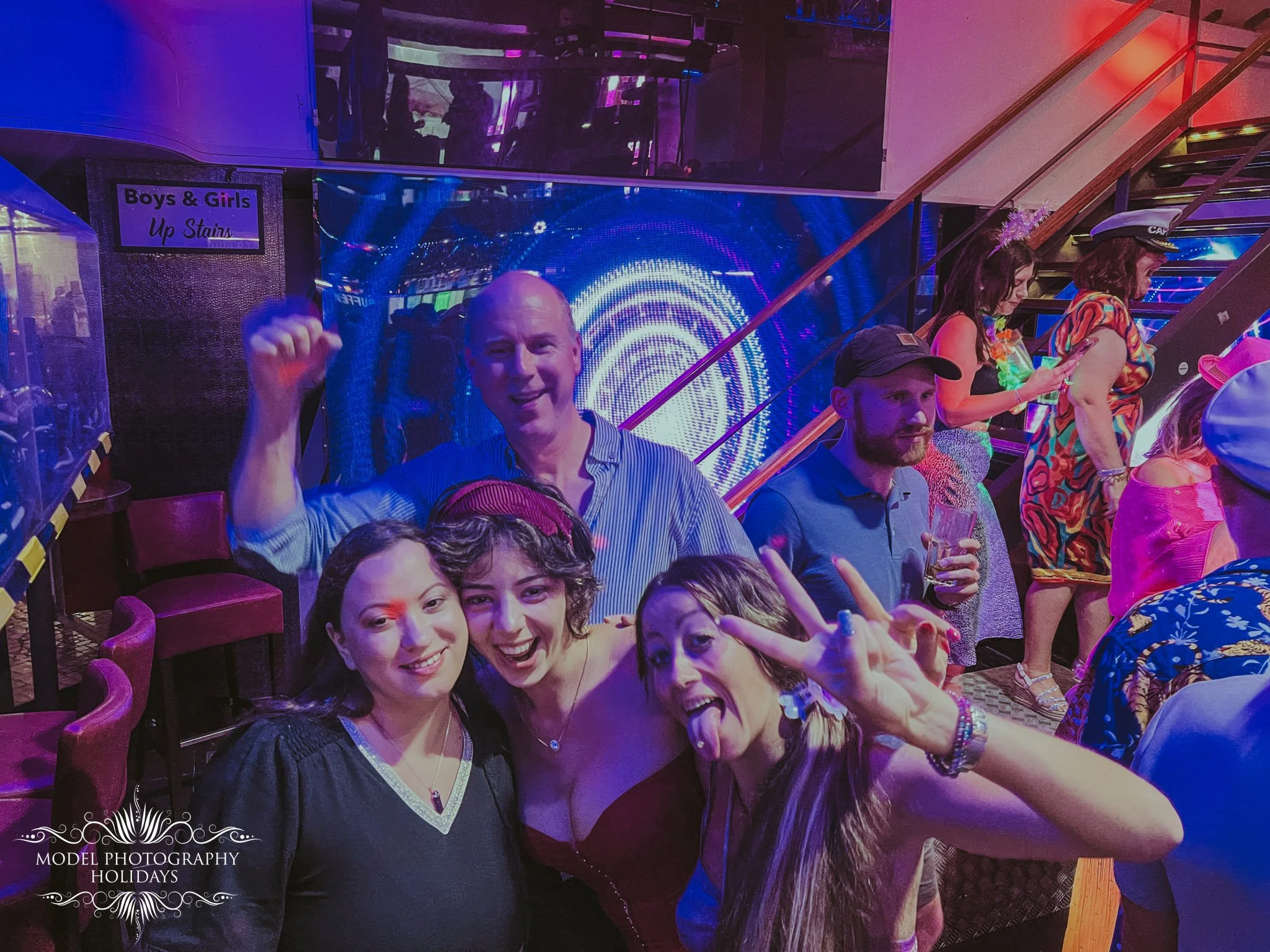 Group of people at a lively party with colorful lighting, some in costumes. A man in striped shirt waves at the camera, surrounded by smiling women. A staircase is visible in the background.