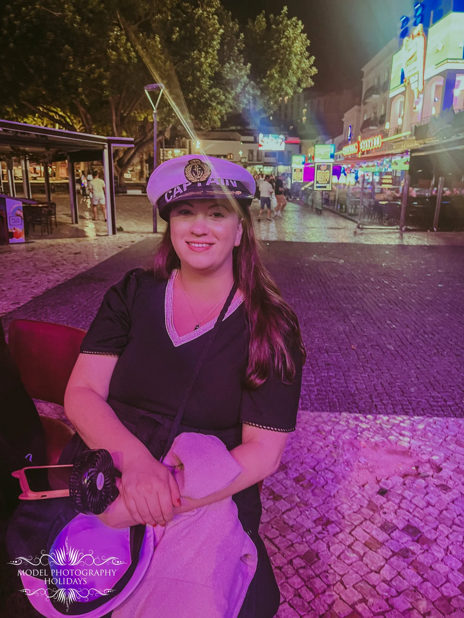 A young woman with long brown hair and light skin smiling at night in an illuminated street with neon signs and gaming machines in the background, wearing a captain's hat and a black top with a light-colored shirt underneath.