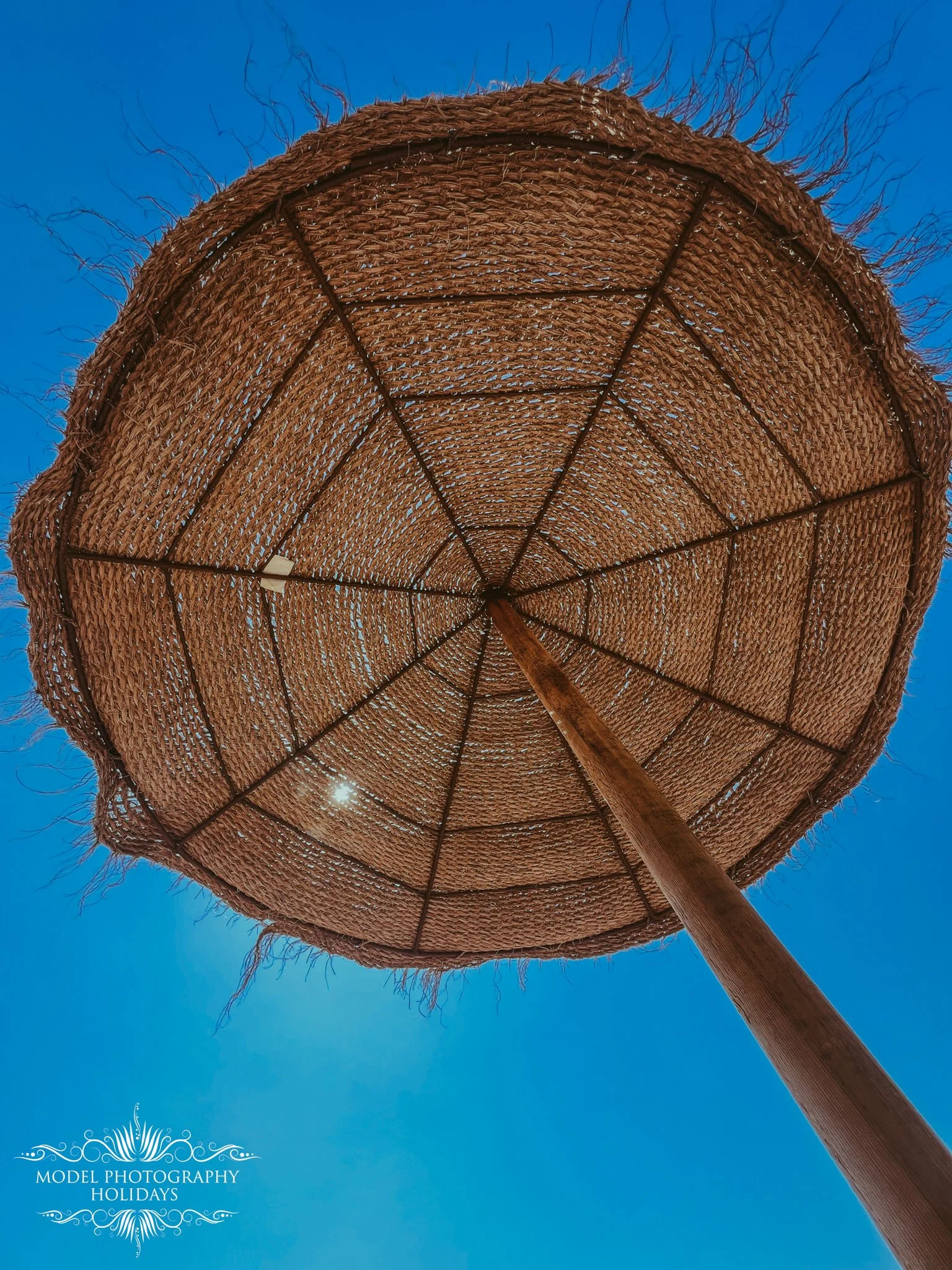 A view looking up at a large woven straw beach umbrella with a wooden pole against a bright blue sky.