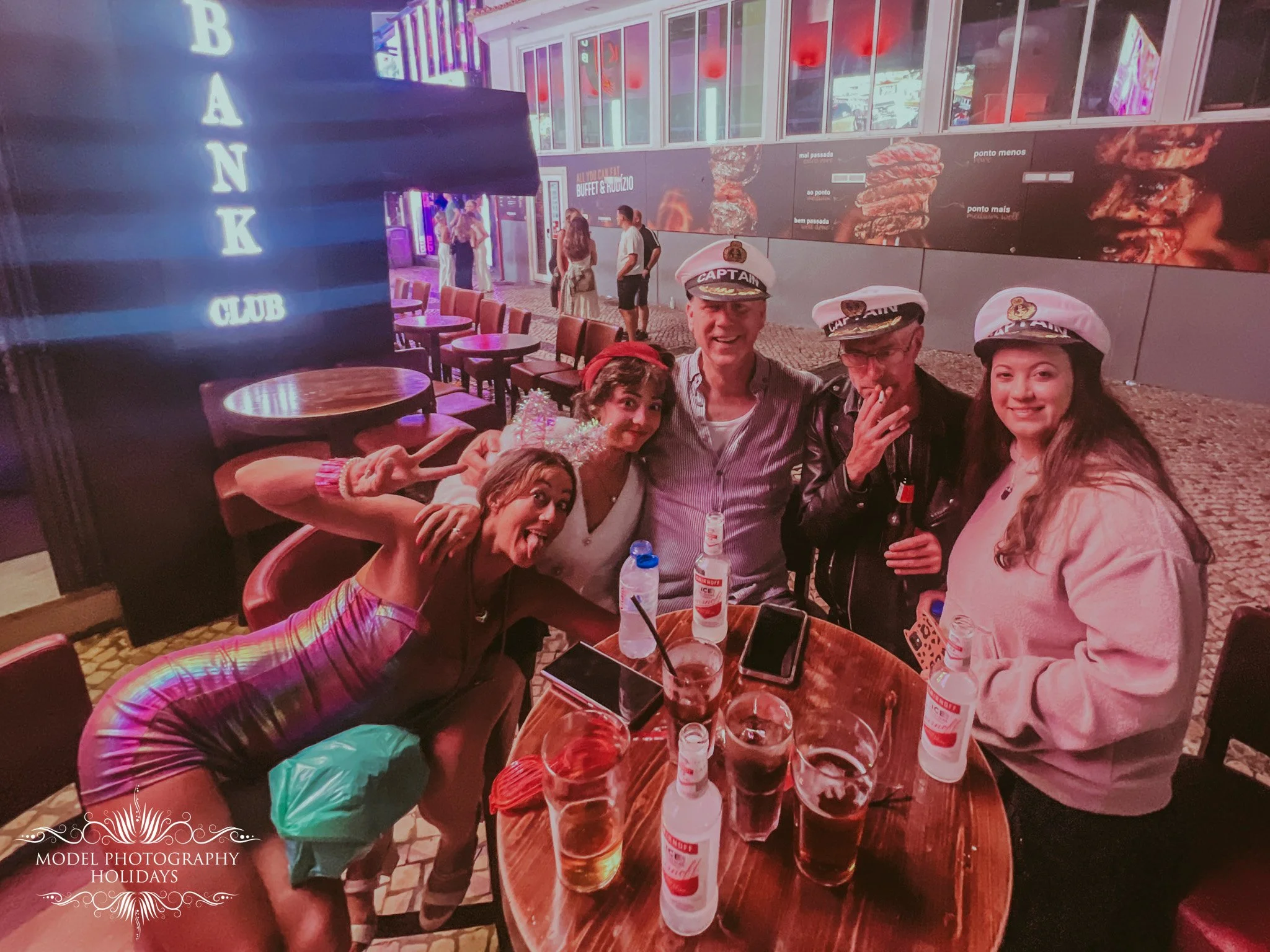 Group of five people, three women and two men, posing for a photo at an outdoor restaurant at night. They are smiling, some making peace signs, and wearing casual clothing including captain hats. The table in front has drinks and bottles. In the back