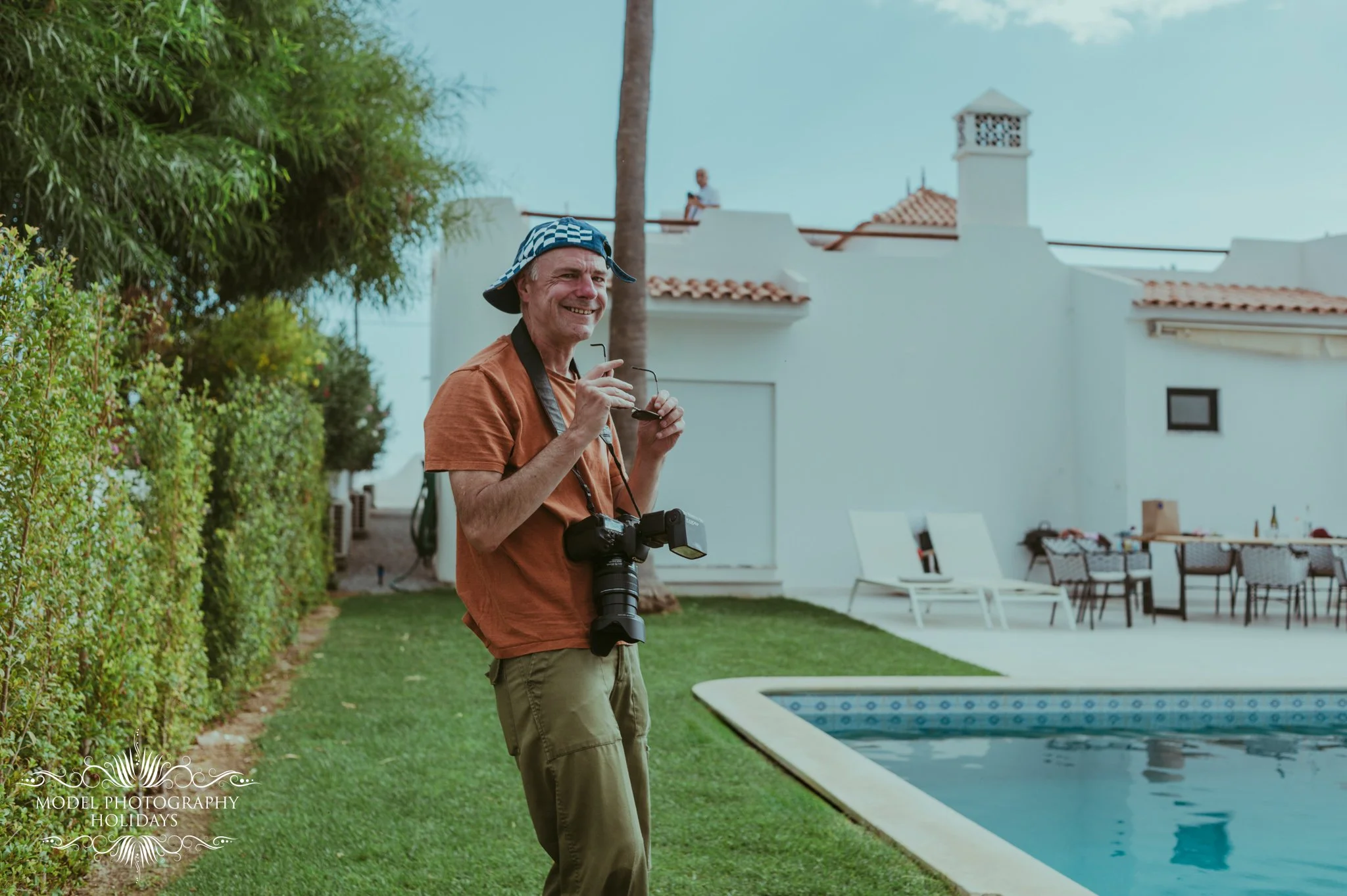 Man holding a camera and sunglasses near a backyard swimming pool, with white house and outdoor furniture in the background