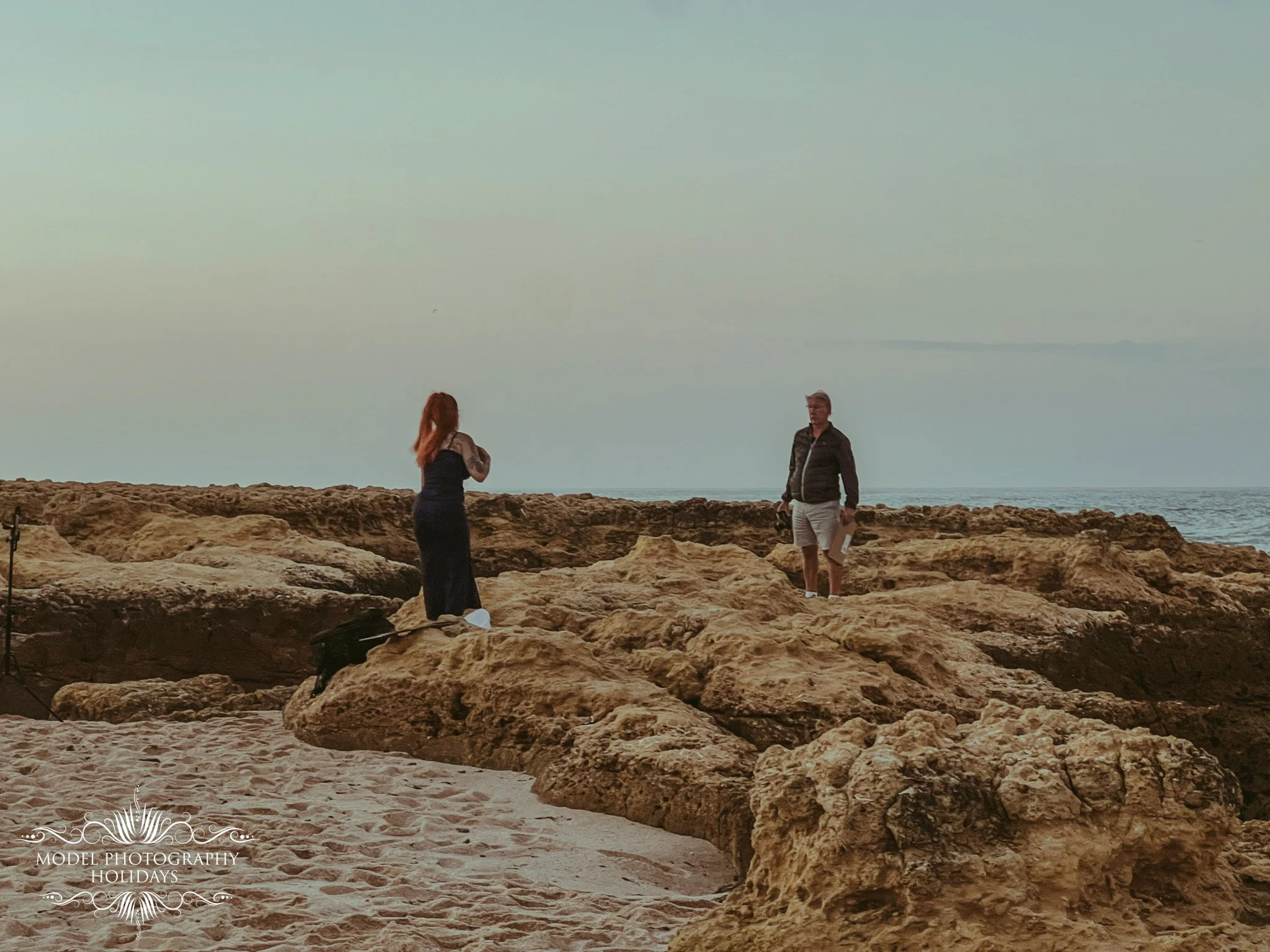 Two people standing on rocky shoreline near the ocean with an overcast sky and distant landmass visible.