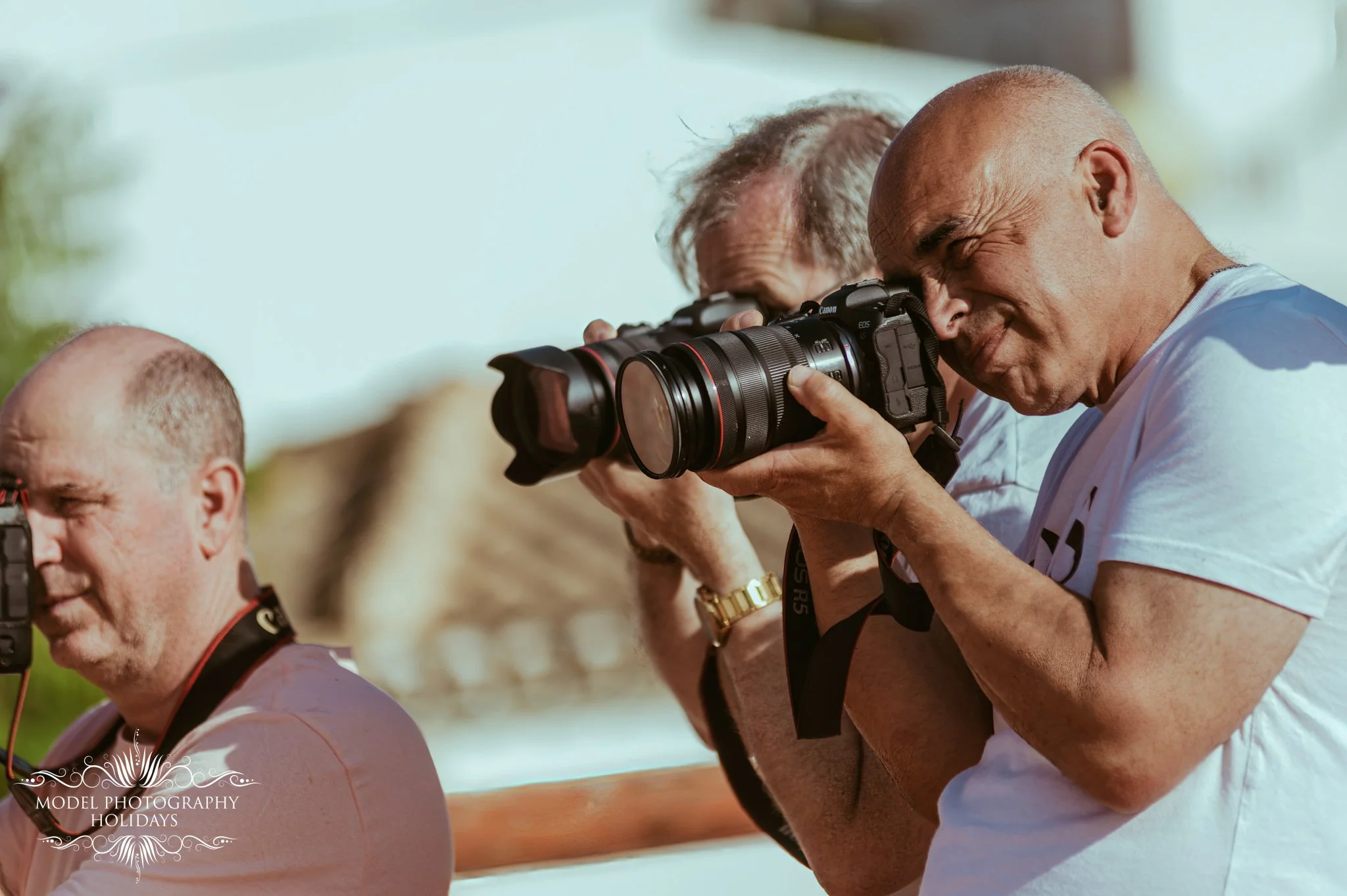 A group of men, some wearing glasses, taking photos using professional cameras outdoors on a sunny day. The man in the foreground is smiling and looking through a Canon EOS camera.