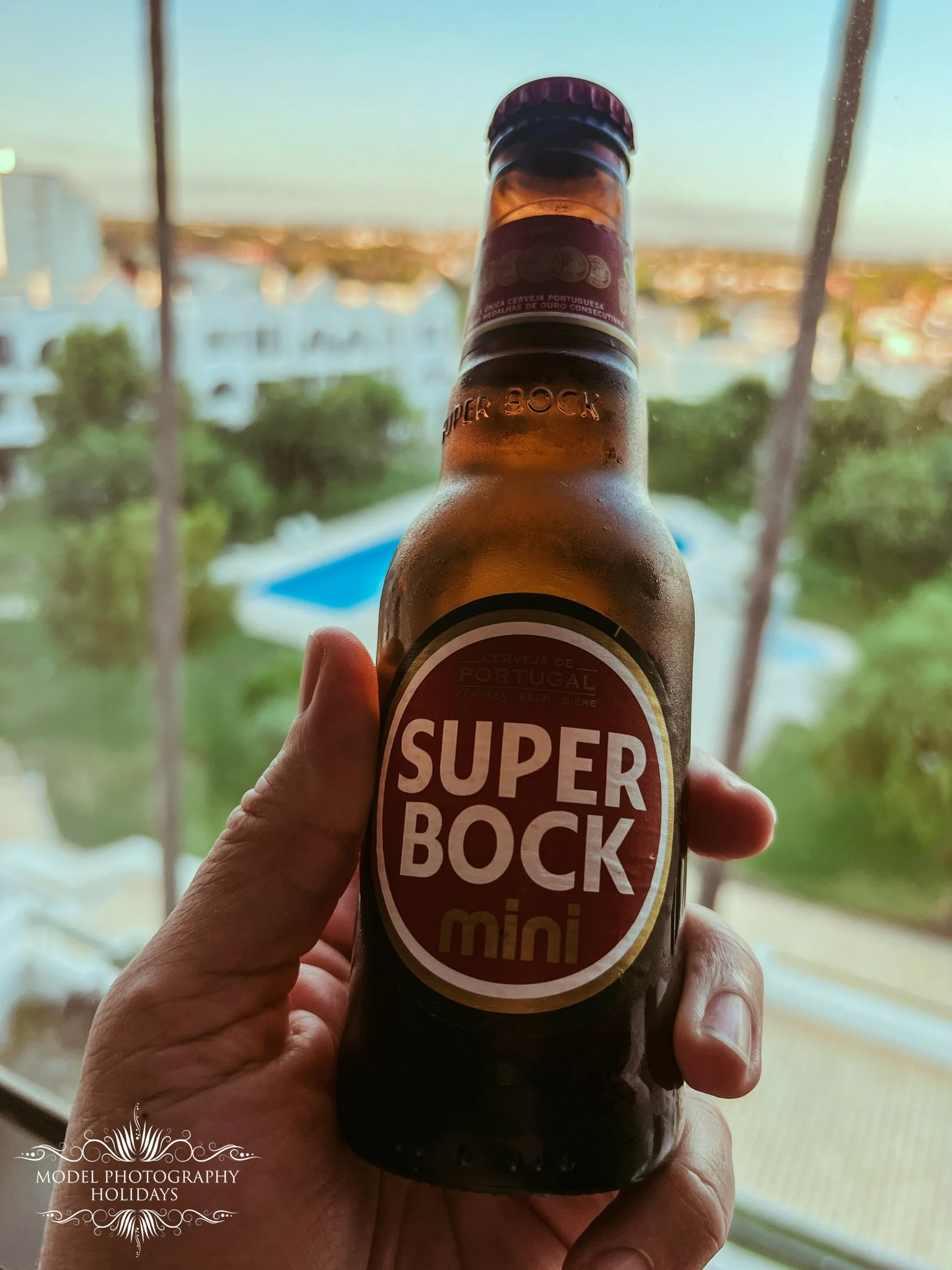 Close-up of a hand holding a small bottle of beer with a label that reads 'Super Bock Mini', with a blurred outdoor background visible through a window.