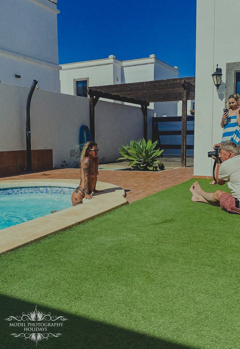 Outdoor photoshoot with a person in sunglasses near a pool, surrounded by photographers and a plant, against a backdrop of white buildings.
