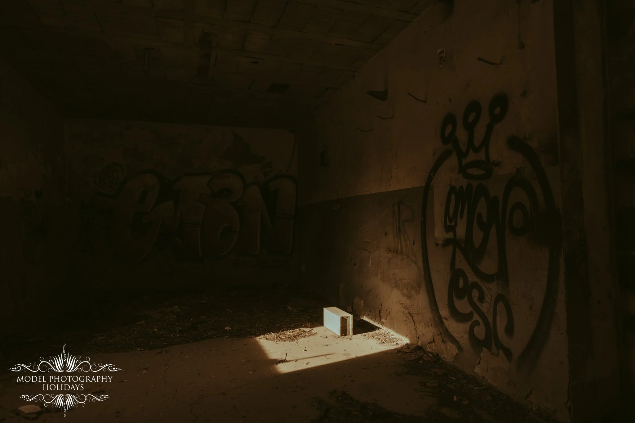 An abandoned building with graffiti on the walls and a floor covered in dirt and debris. A single concrete block is placed on the ground near a beam of sunlight coming through an opening.