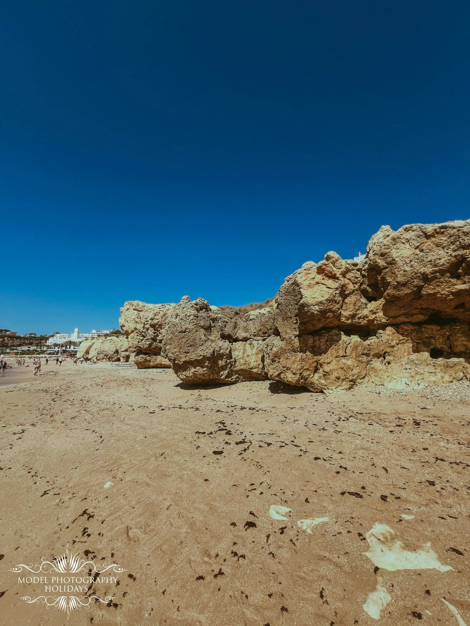 A sandy beach with large rocks near the shore, with a clear blue sky above and some buildings visible in the distance.