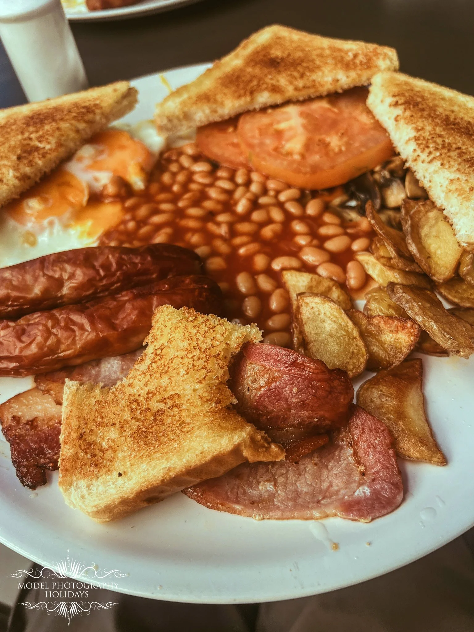 A breakfast plate with toast, baked beans, fried eggs, sausage, and bacon.