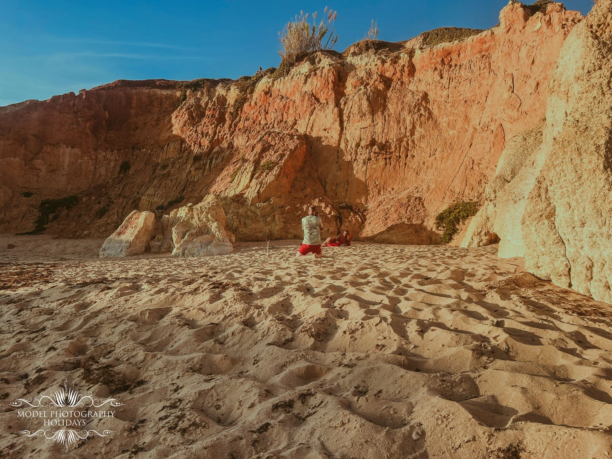 A person taking a photo of another person lying on the sandy beach next to large rock formations and reddish-orange cliffs on a clear day.