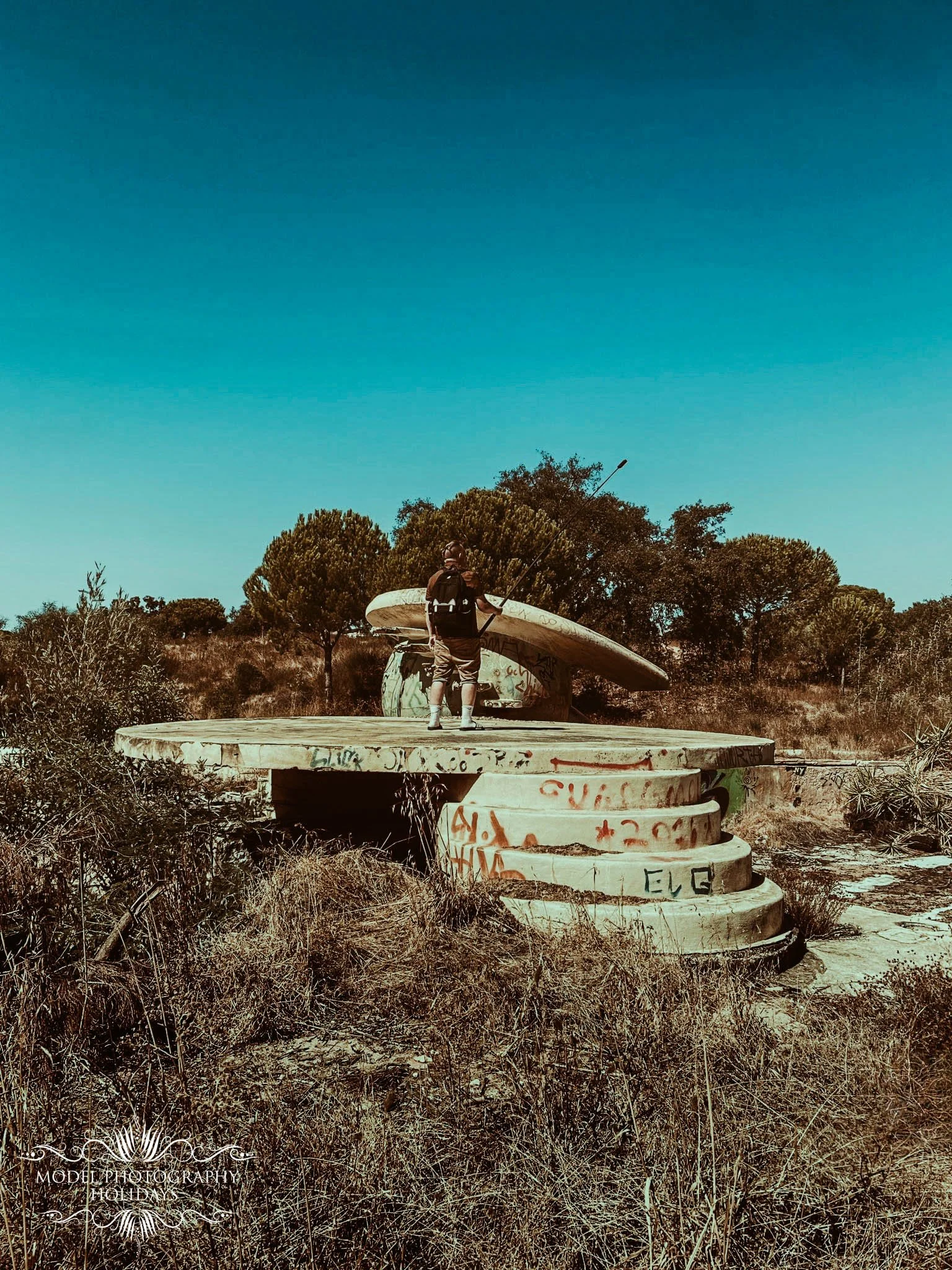 A person with a backpack holding a paddle at an abandoned boat graveyard with graffiti on the boats, surrounded by dry vegetation and trees under a clear blue sky.