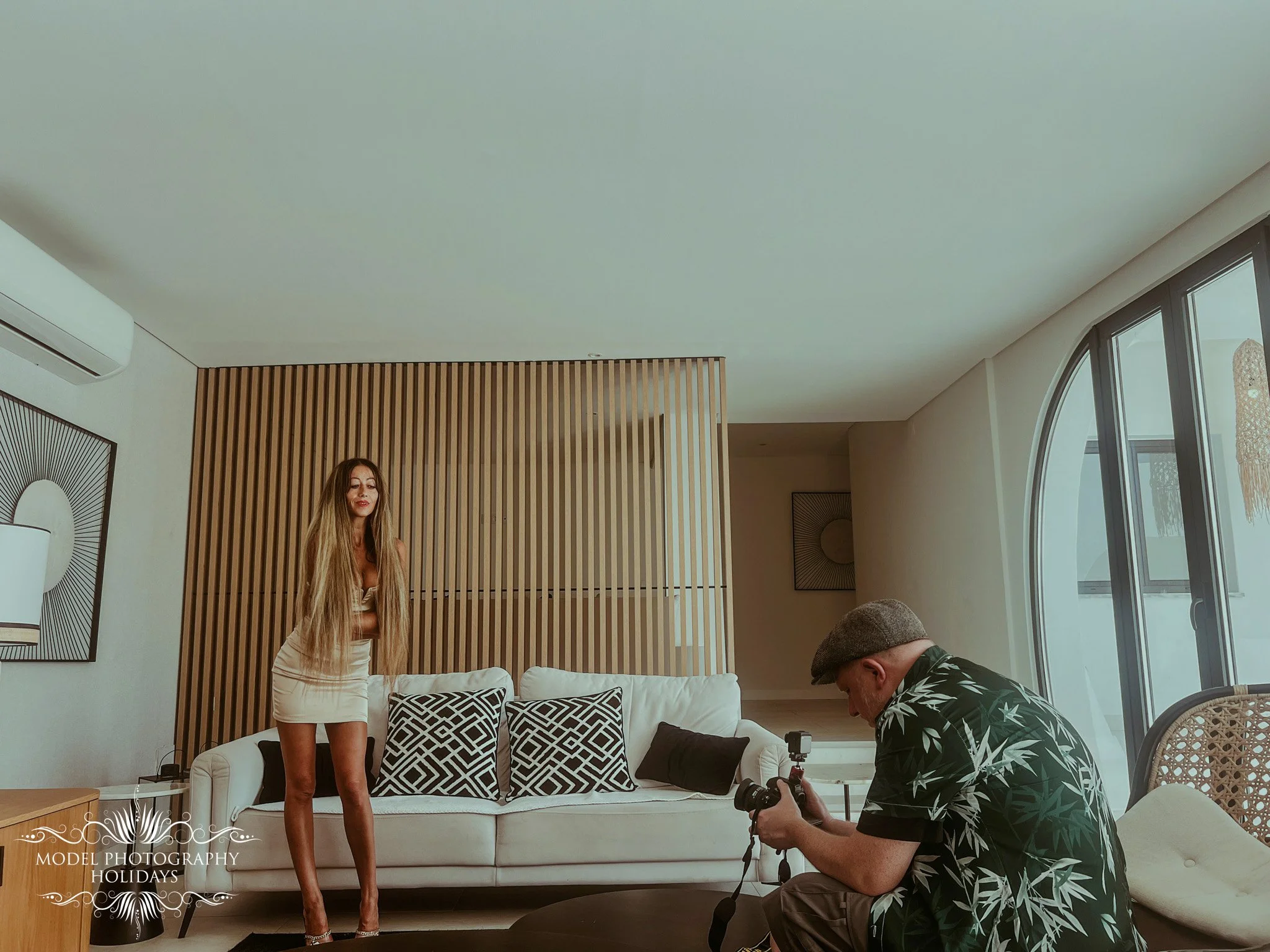 A woman with long blond hair and a beige dress standing in a modern living room, with a man in a black floral shirt kneeling and adjusting a camera, a white couch with black and white patterned pillows, and decorative artwork on the wall.