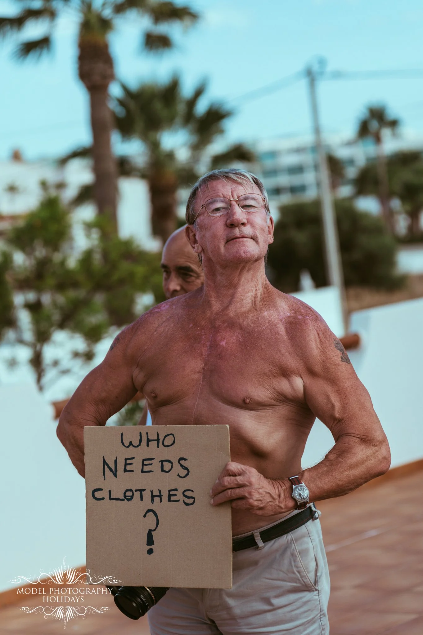 A shirtless elderly man with glasses holding a cardboard sign that says "Who needs clothes?". Behind him, another person is partially visible, and there are palm trees, a building, and power lines in the background.