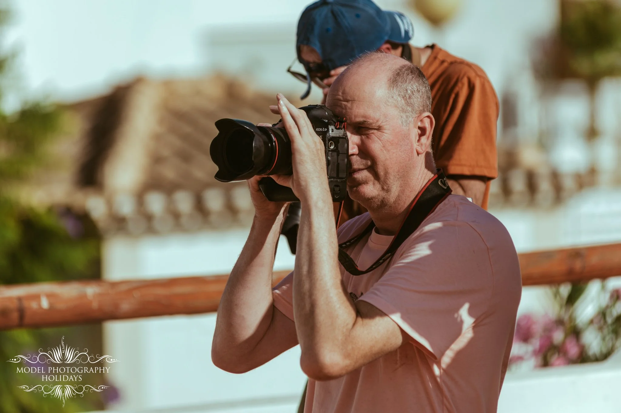 A man with a bald head and beige shirt taking a photo with a DSLR camera, while a young person with glasses, a blue cap, and a brown shirt observes behind him. They are outdoors with a wooden fence and trees in the background.