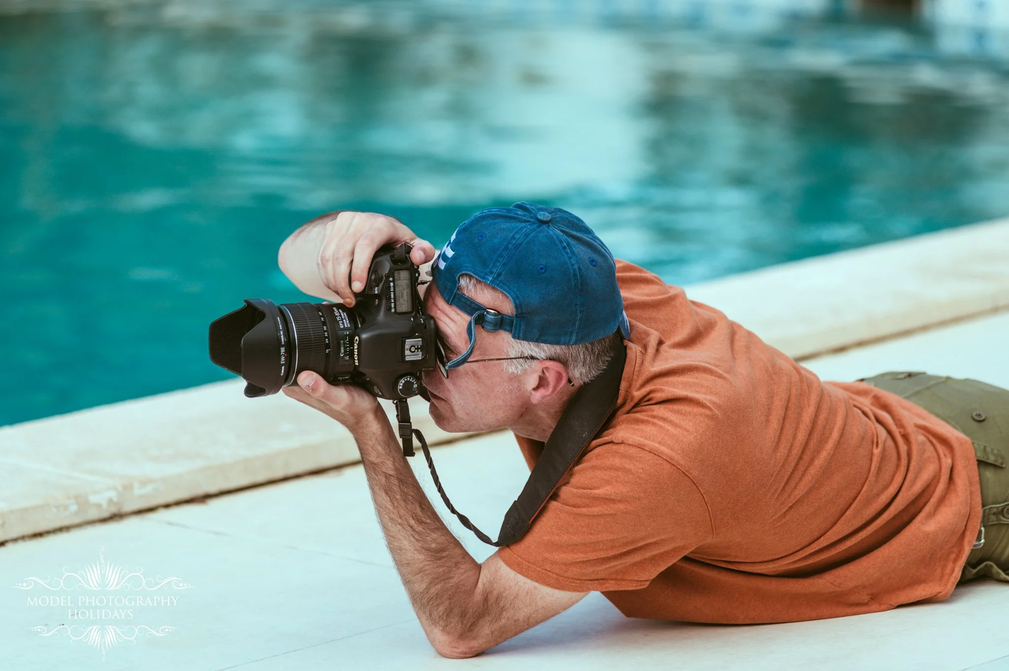 A man lying on the ground with a camera, aiming through the viewfinder and taking a photograph by a swimming pool.